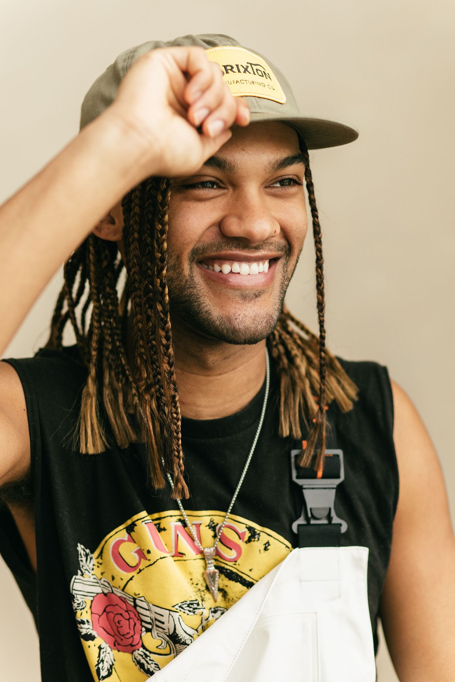 Smiling Brandon Cameron with dreadlocks wearing a black tank top and baseball cap adjusts their hat while posing for a photo.
