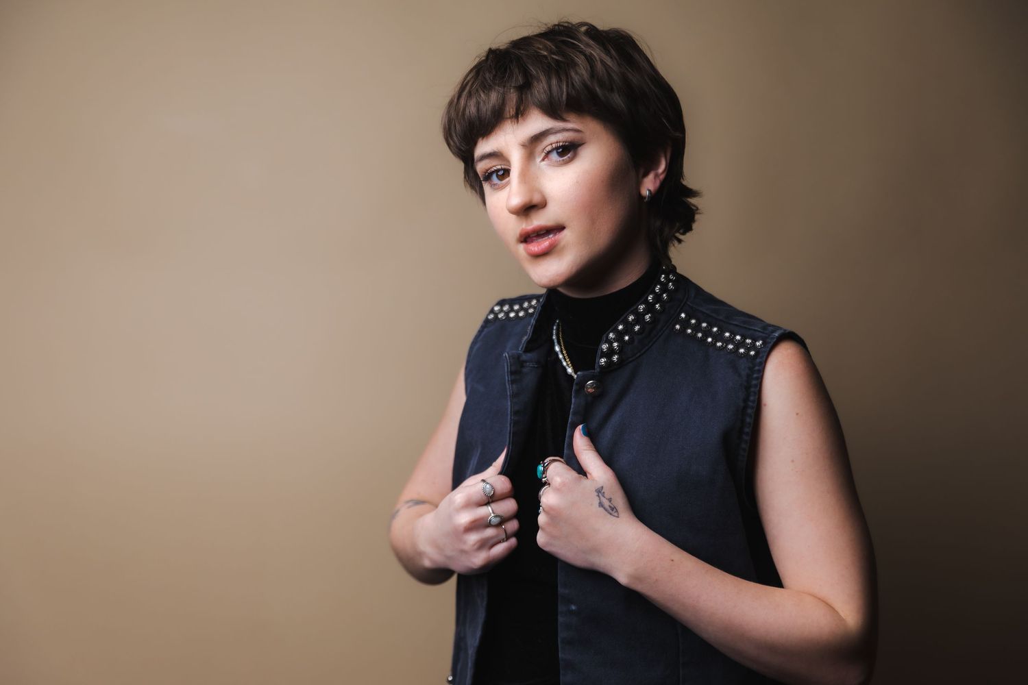 Studio portrait of a person wearing a black studded vest against a neutral beige background.