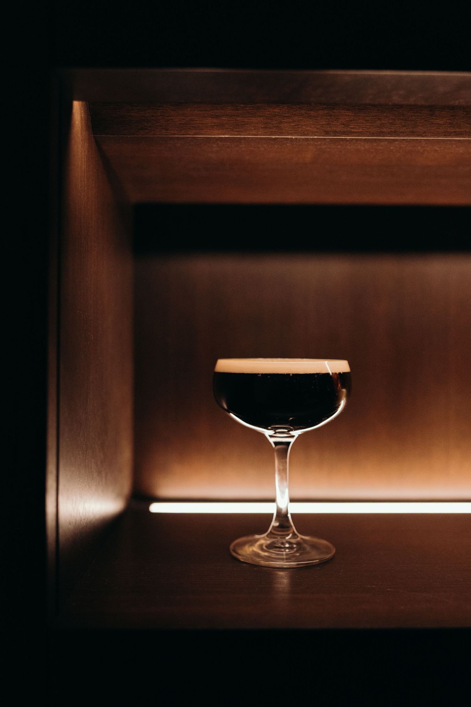A dark beer with creamy foam head sits in an elegant cocktail glass against a moody wooden backdrop at the Algonquin Hotel
