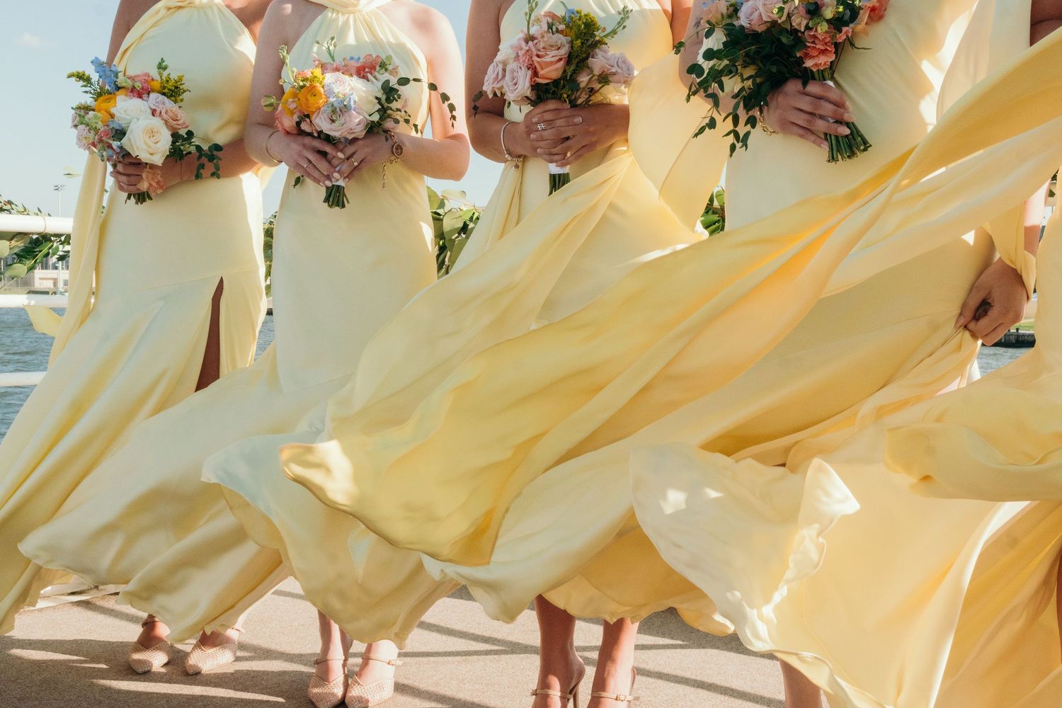 Bridesmaids in pale yellow dresses pose together holding bouquets at an outdoor wedding.