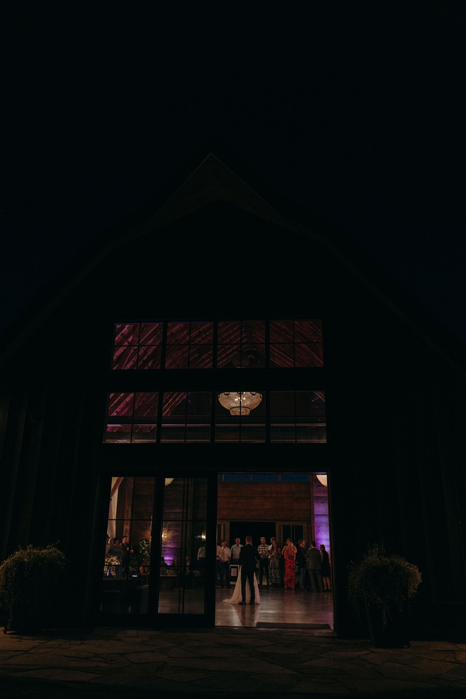 Dark barn venue lit with warm ambient lighting showing guests through large windows at night.