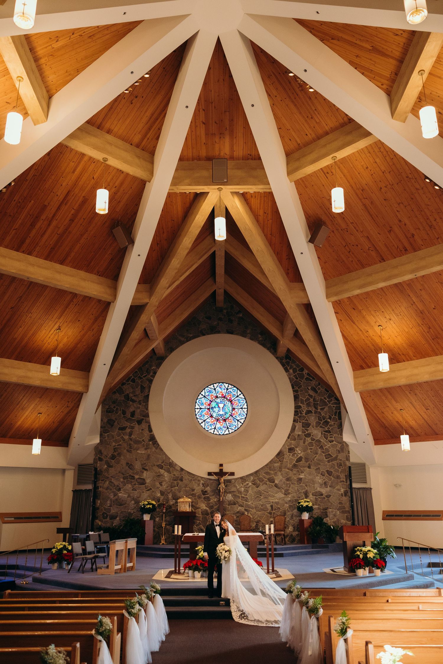 Interior of modern church with vaulted wooden ceiling, stone wall, circular stained glass window and hanging pendant lights.