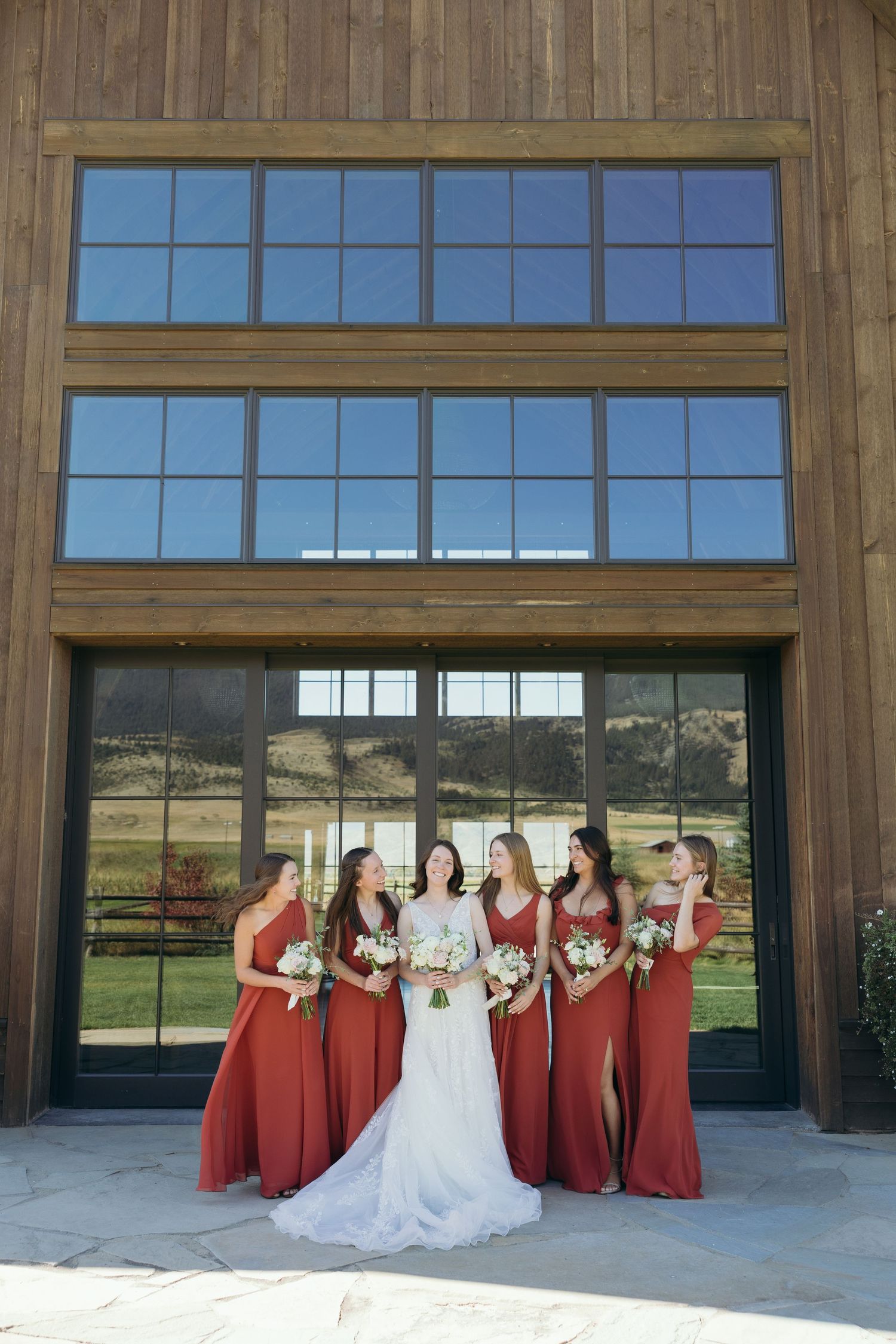 Wedding party poses in front of large barn windows with mountain scenery visible.