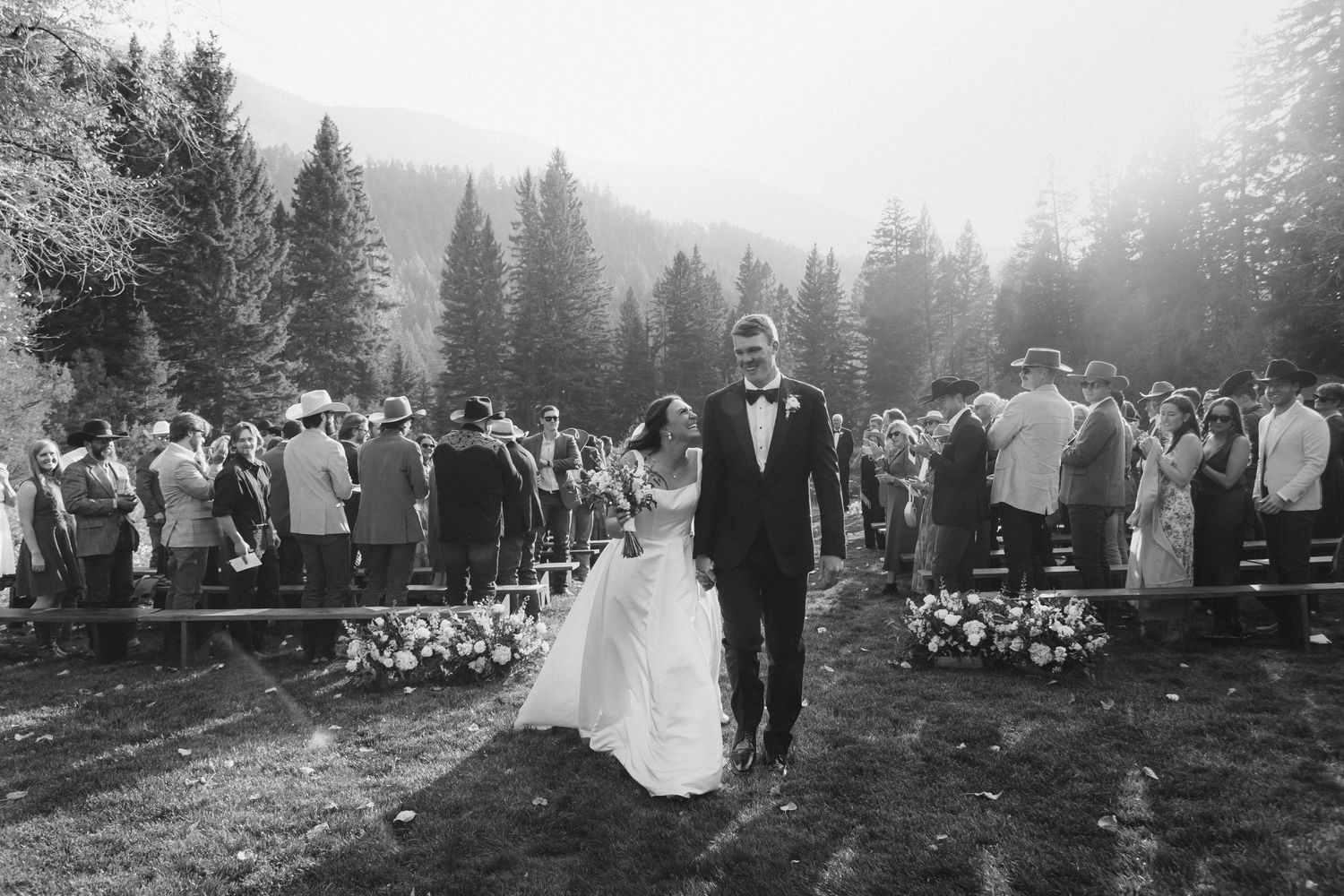 A black and white wedding ceremony photo with guests gathered among tall pine trees.