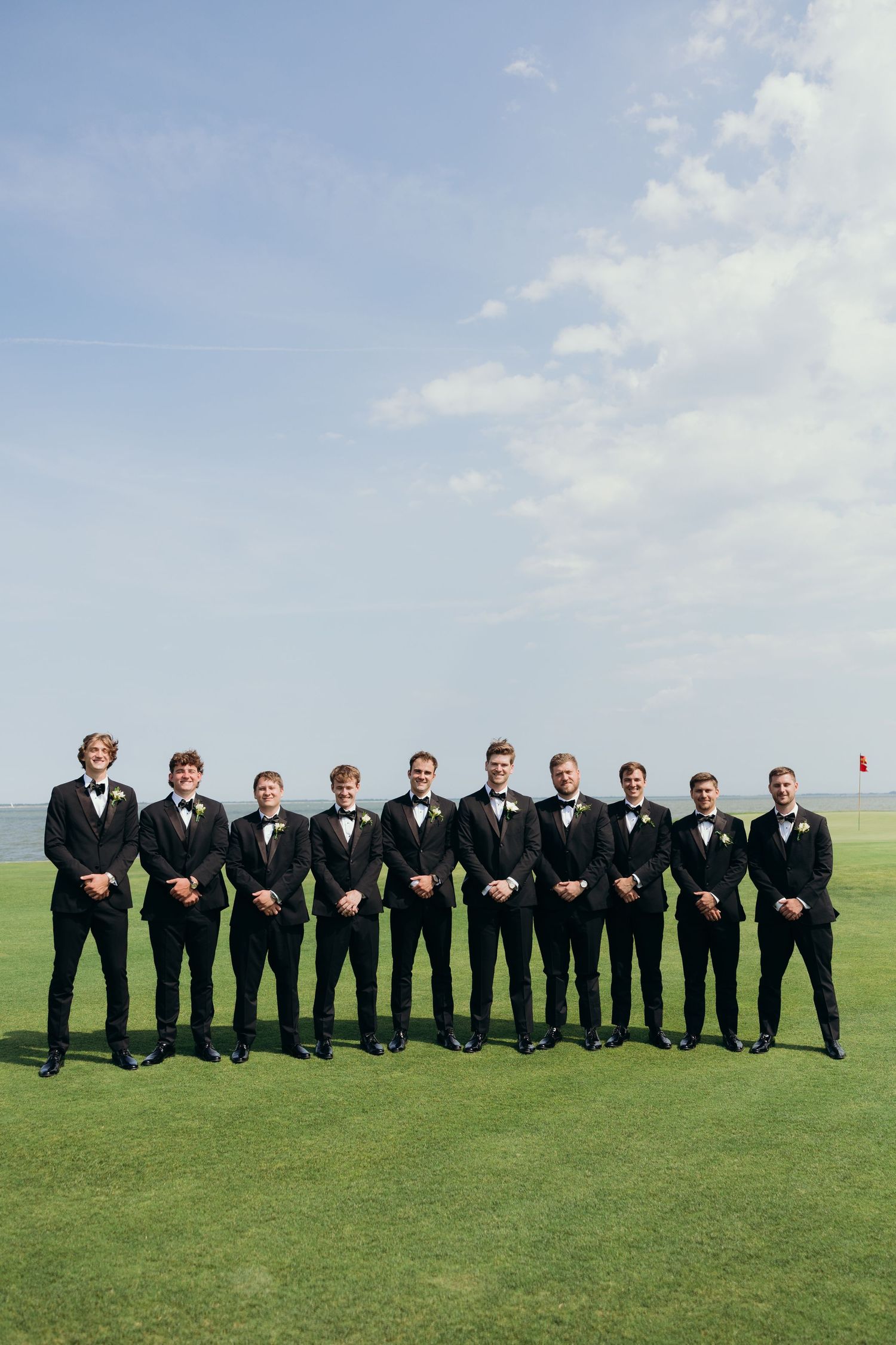 Groomsmen in black suits line up on a green hilltop for a formal wedding photo.