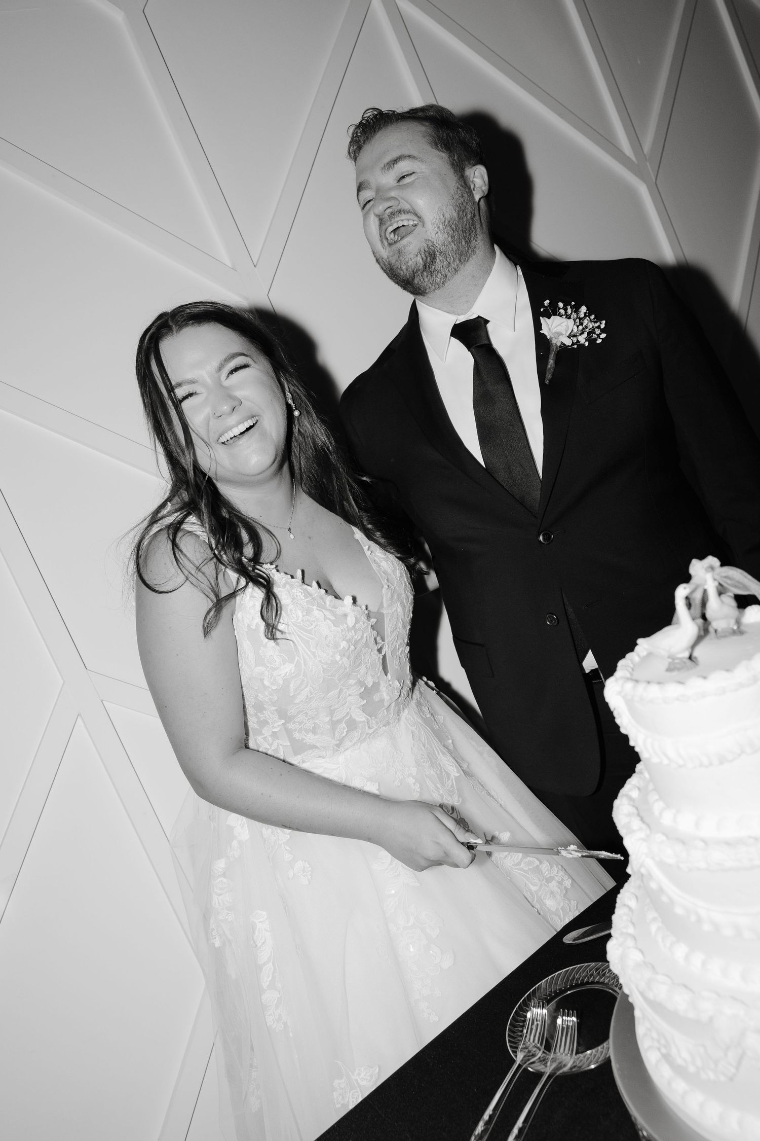 Joyful wedding couple laughing while cutting their white tiered cake together.