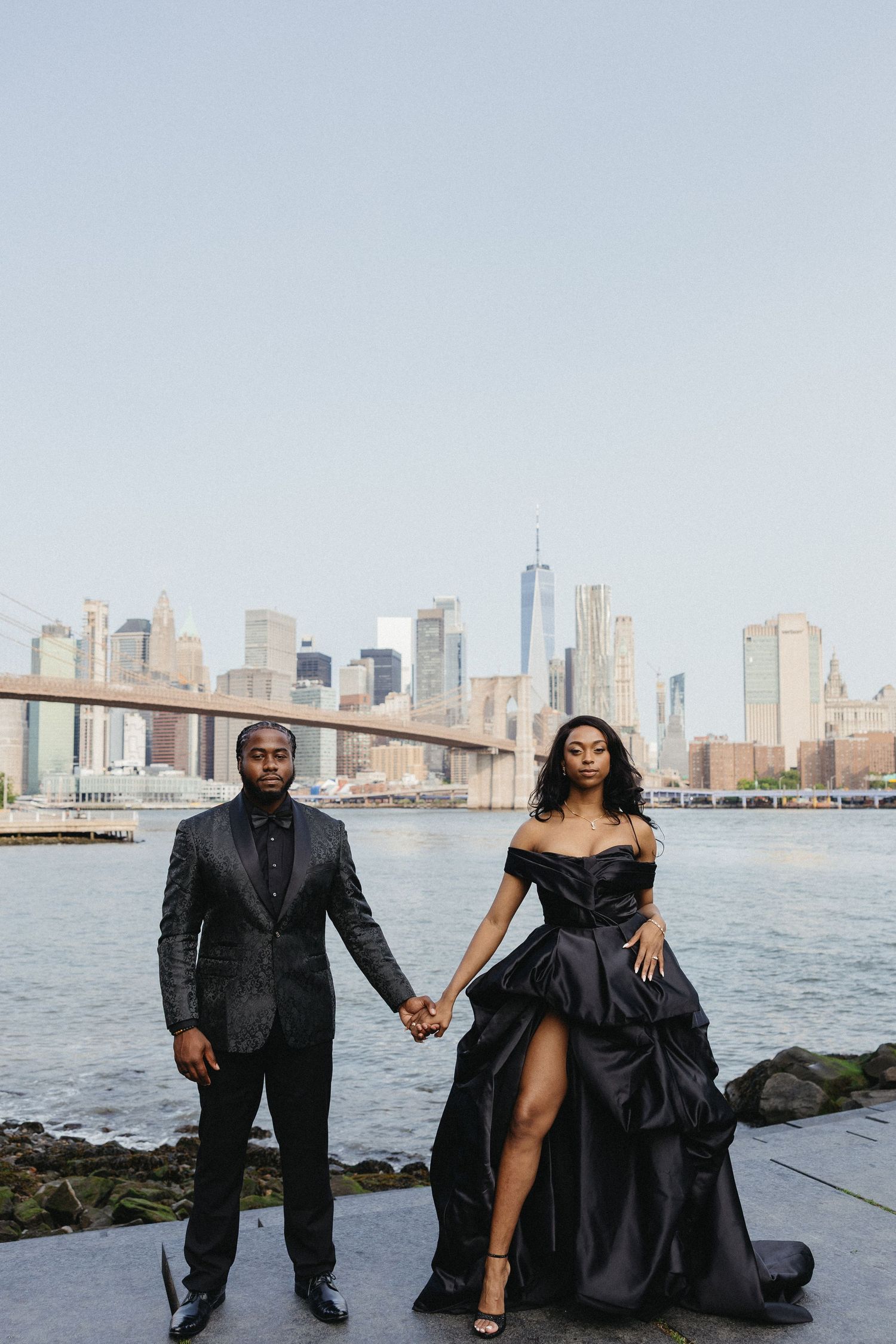 Elegant couple in formal black attire poses along waterfront with city skyline view.
