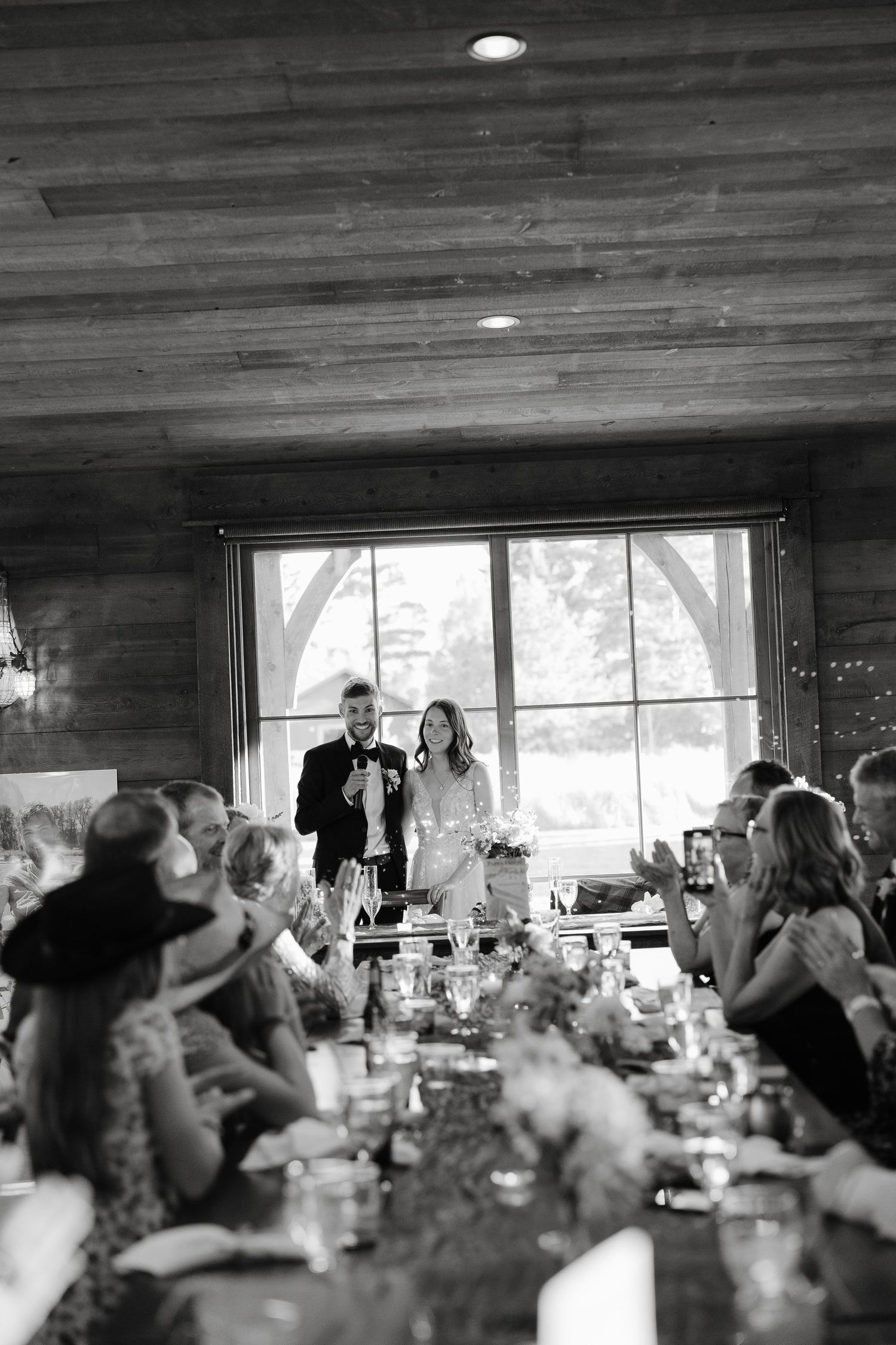 Black and white photo of newlyweds giving wedding reception speech at rustic venue.
