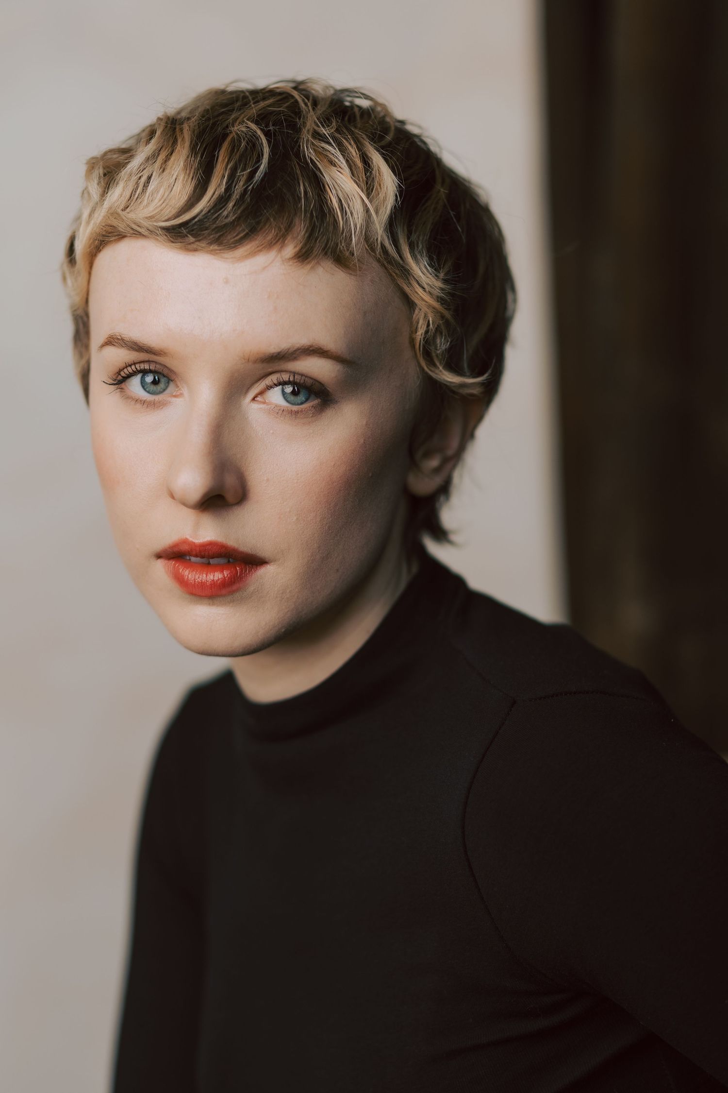 Portrait photo with short blonde wavy hair, red lipstick, and black turtleneck against neutral background.