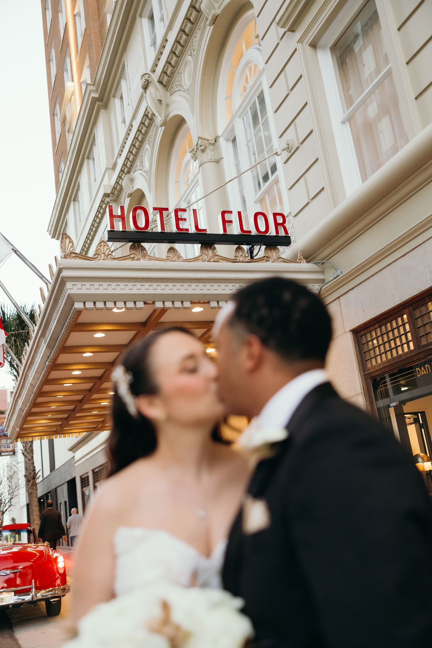 Romantic wedding moment captured under the vintage Hotel Flor sign with classic car in background.