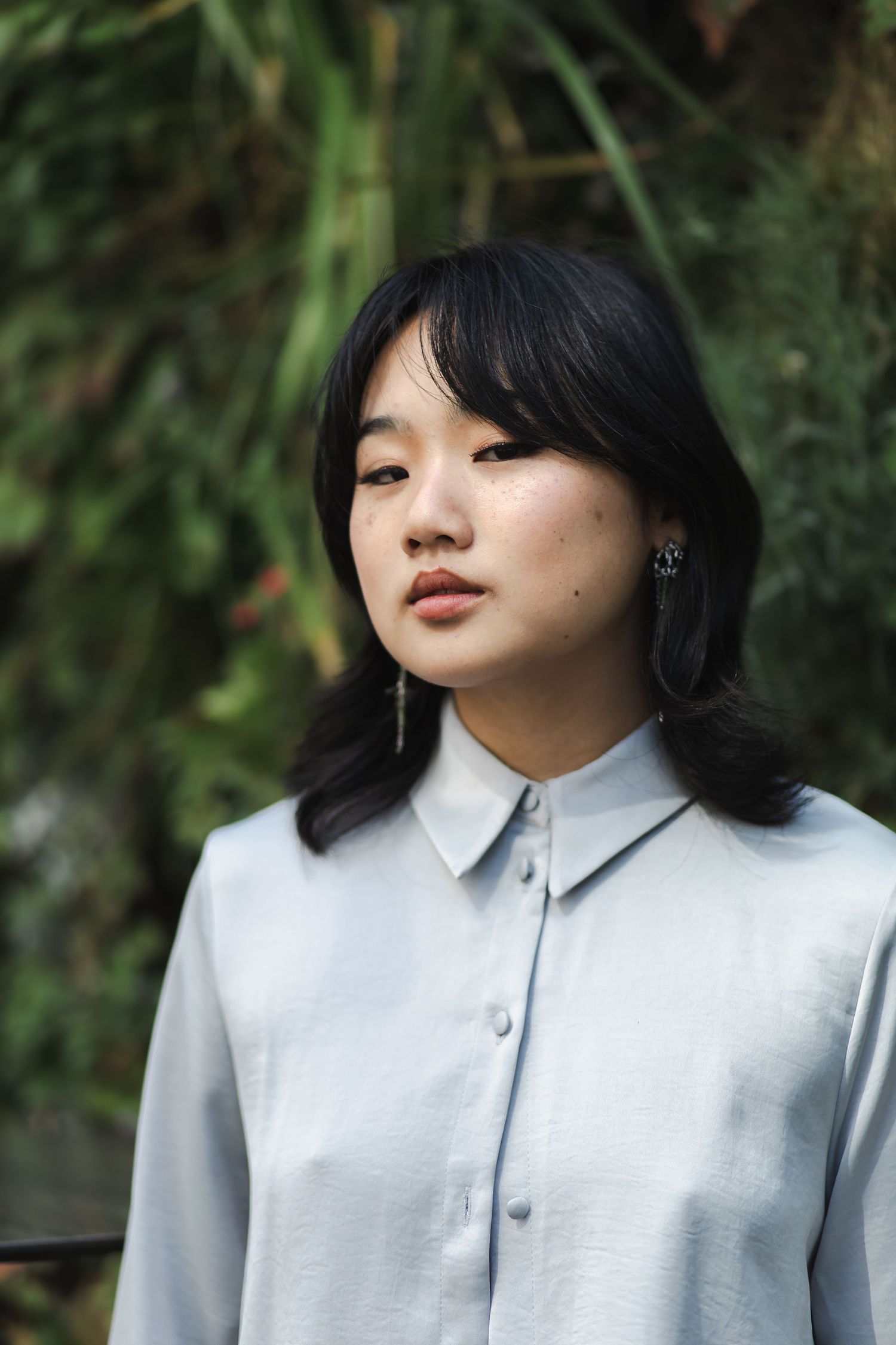 Young Asian woman in light blue button-up shirt with dangling earrings, posing outdoors with green foliage background.