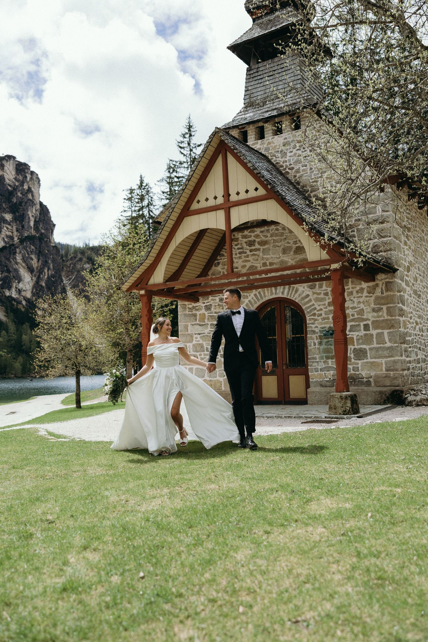 A couple poses outside a rustic stone chapel with a dramatic mountain backdrop in the Alps.