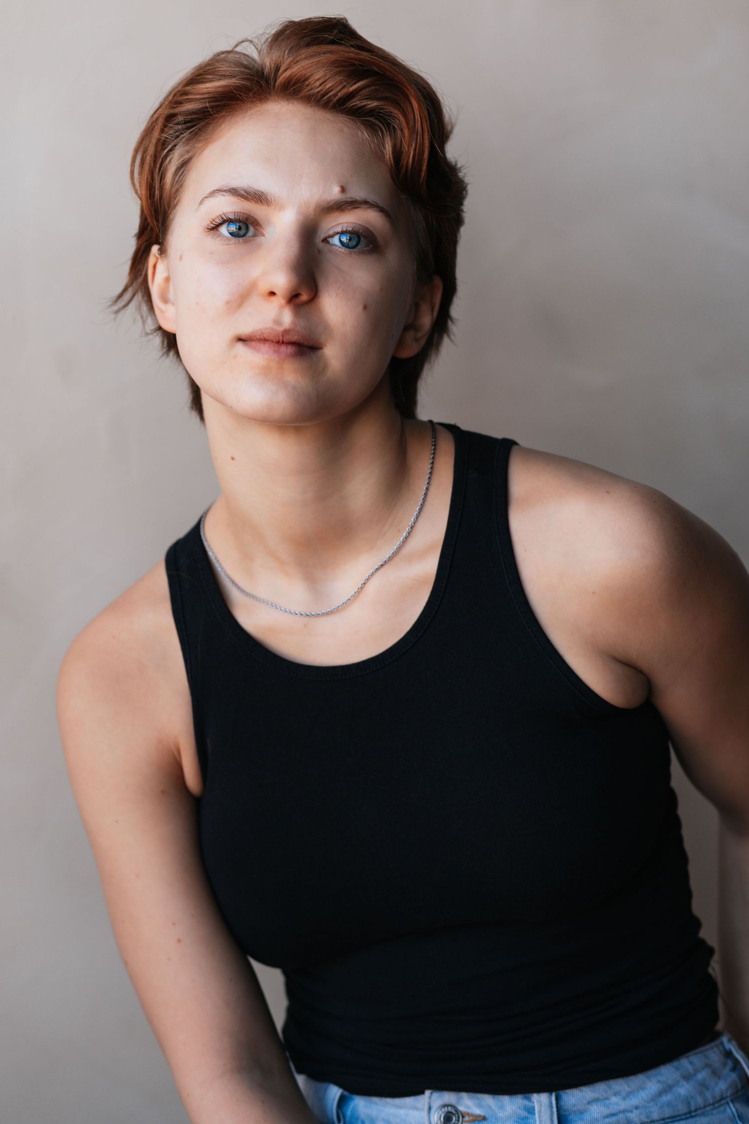 Portrait of someone with short ginger hair wearing a black tank top and jeans against a light gray background.