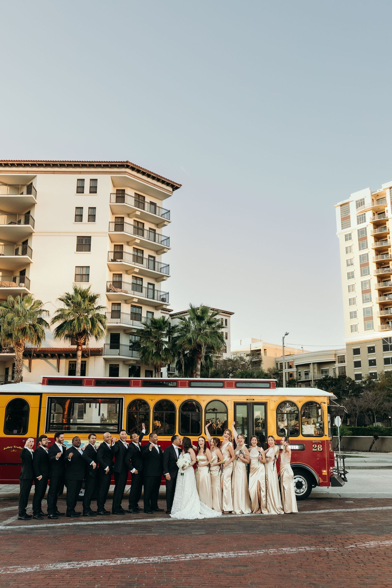 Wedding party poses with vintage trolley bus in front of beachside hotel during golden hour.