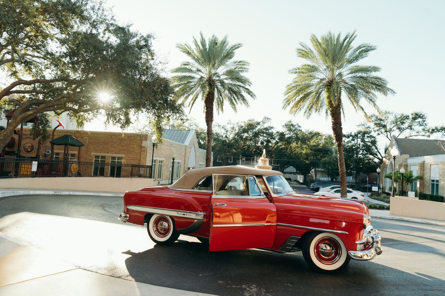 A vintage red car with white wall tires parked on a palm tree-lined street at sunset.