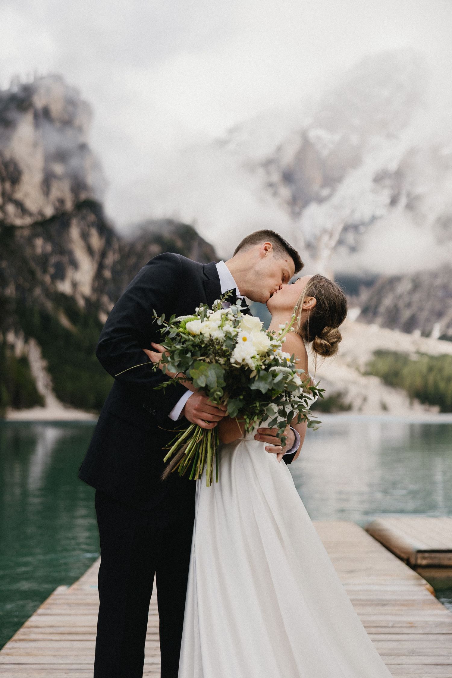Bride and groom sharing a romantic kiss on a dock with mountain scenery and turquoise lake.