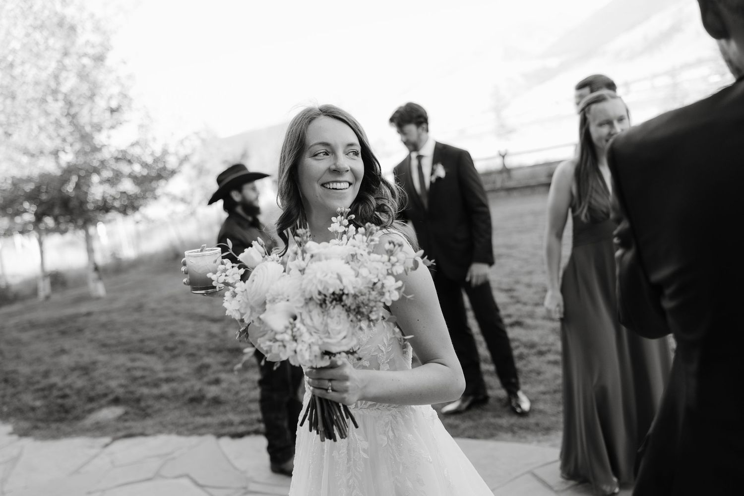 A wedding party poses in front of a rustic barn with mountains in the background.