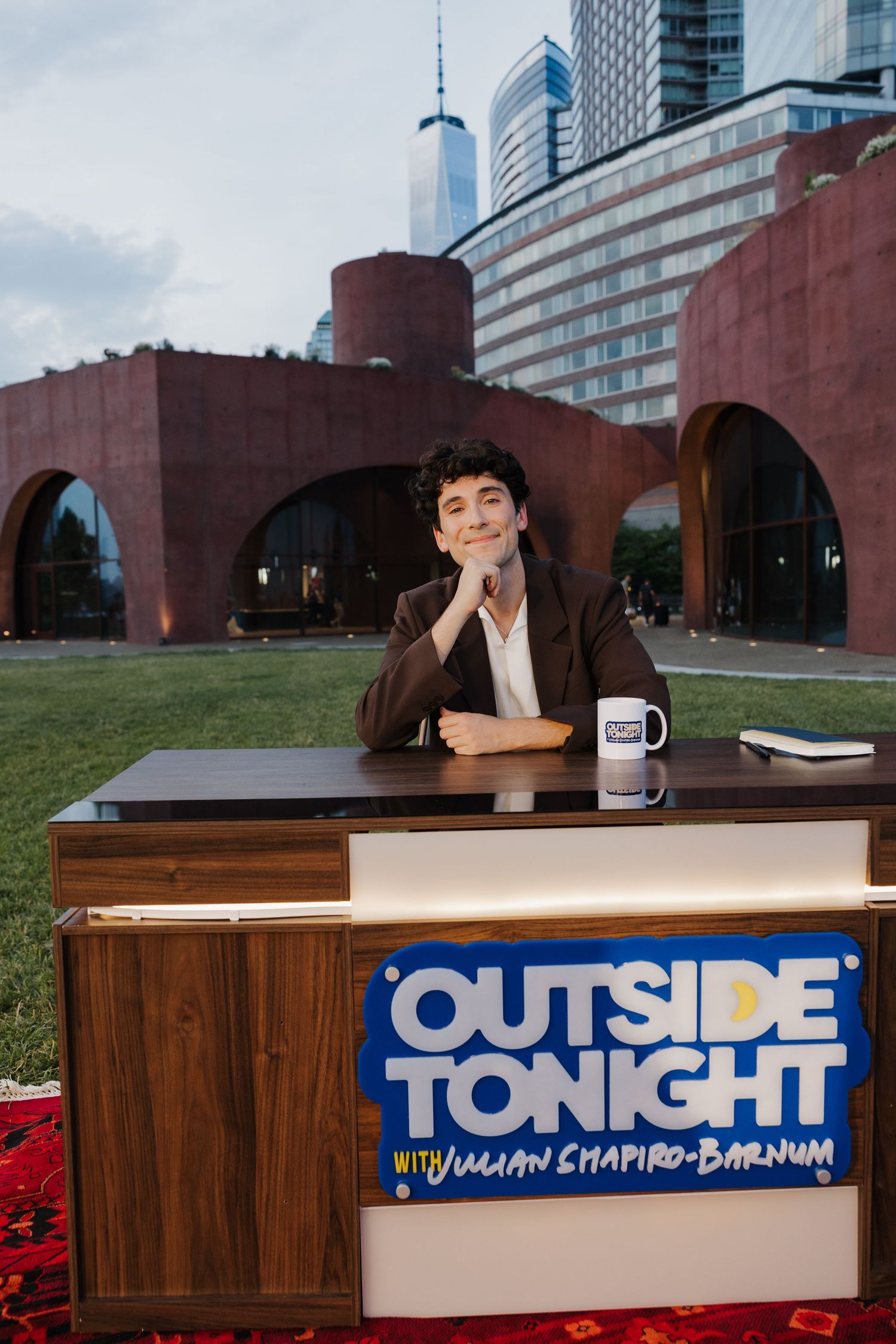 Host Julian Shapiro-Barnum sits at an outdoor desk for Outside Tonight talk show with NYC skyline backdrop.
