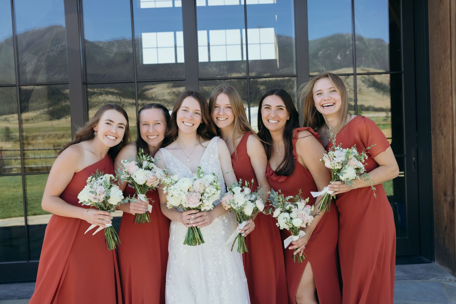 Bridal party in red dresses poses with bouquets against mountain backdrop.