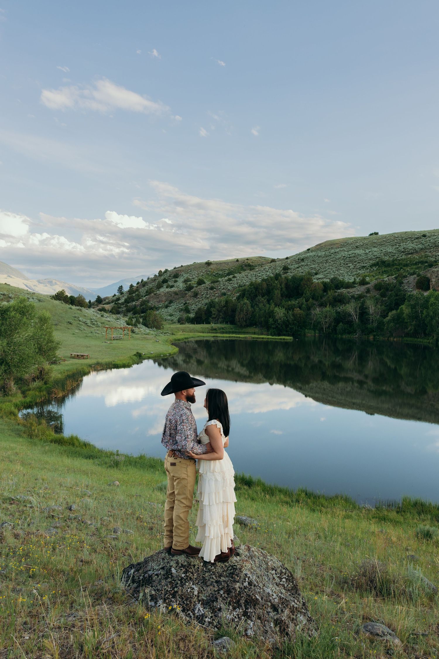 A couple stands together overlooking a serene mountain lake at sunset.