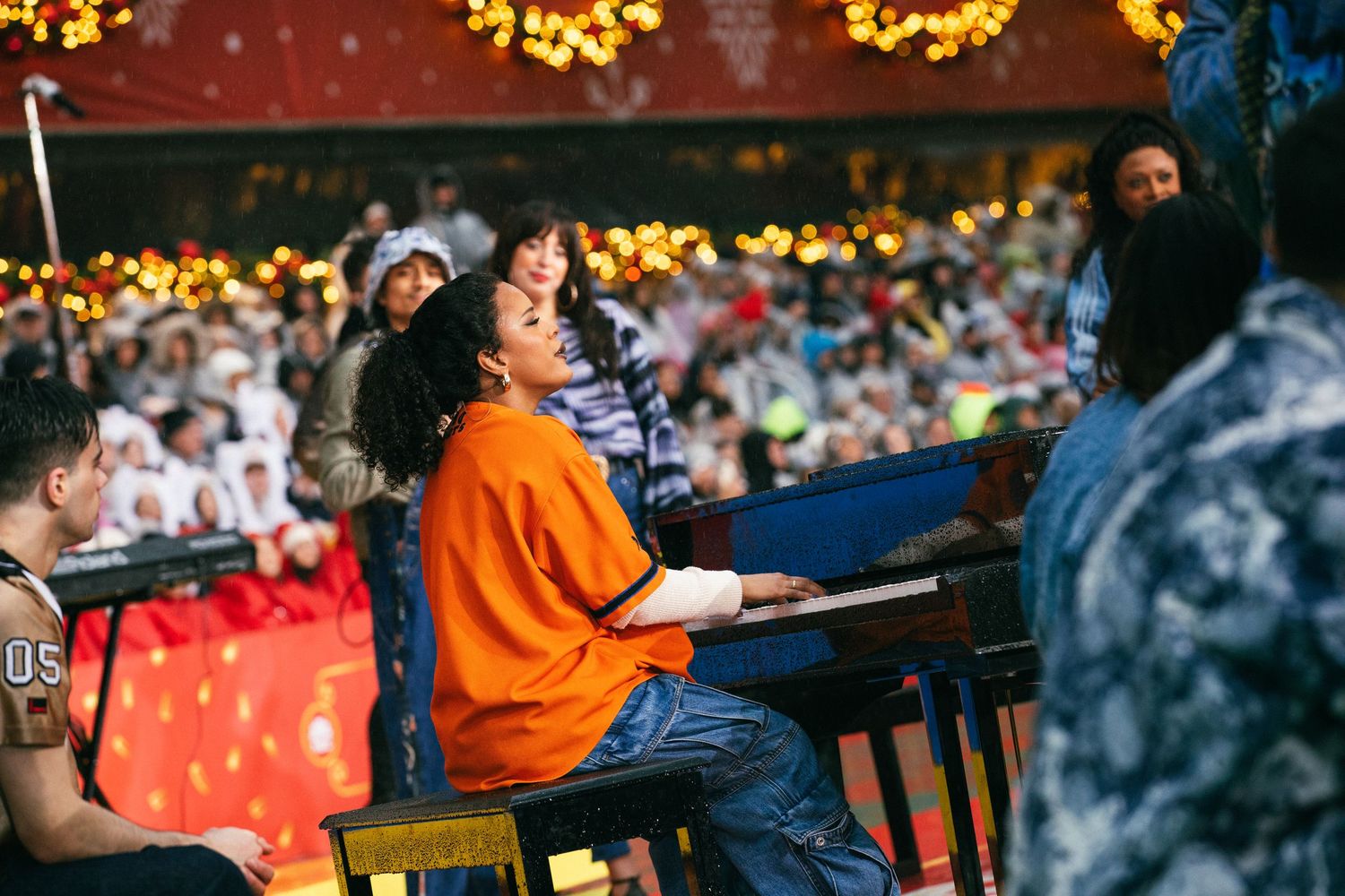 Musician playing piano during outdoor holiday performance with festive lights in background.