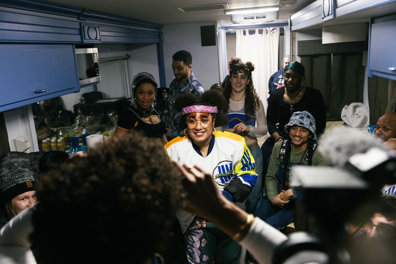 A lively group gathers in a cramped backstage area with colorful outfits and sunglasses.