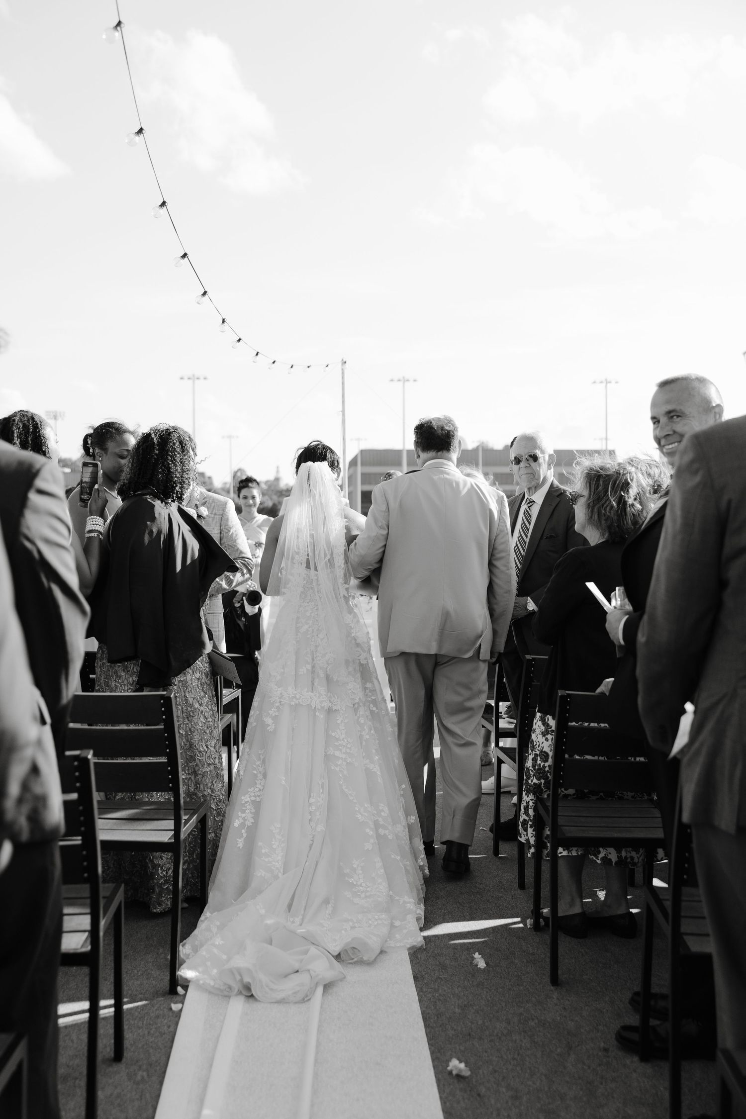 Wedding ceremony guests stand among rows of chairs at an outdoor venue.