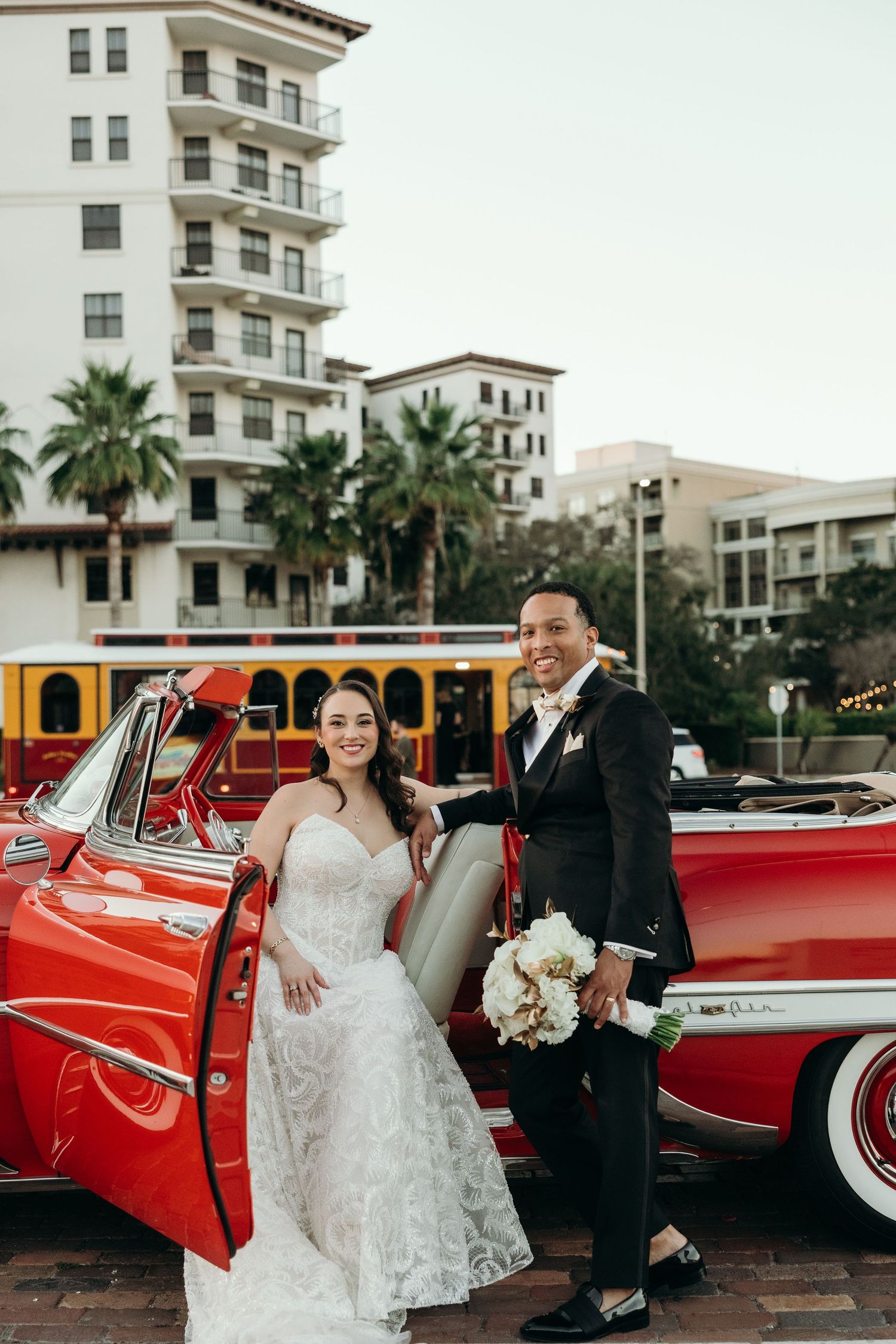 Wedding couple poses with vintage red car in front of Mediterranean style building with palm trees.