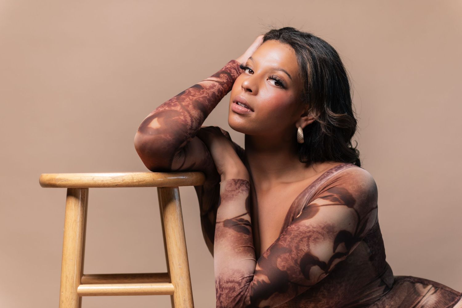 A person in a brown outfit poses on a wooden stool against a neutral backdrop in a professional studio setting.