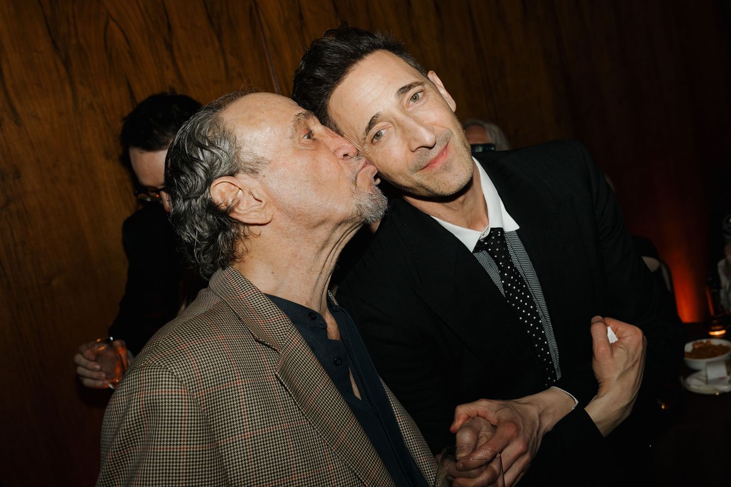 Two men at a social event, one older in a brown jacket leaning in to kiss the cheek of a younger man in a black suit.