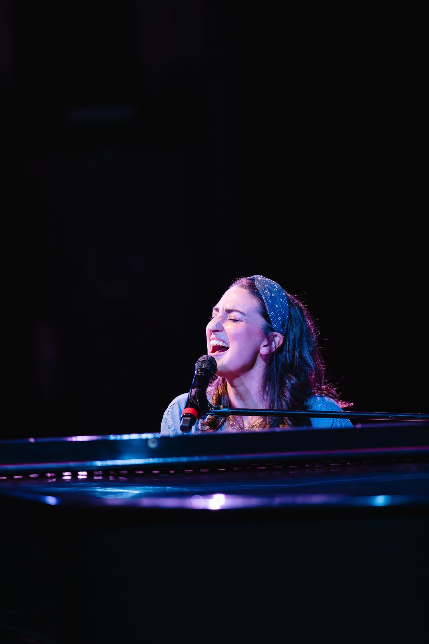 Dramatic stage lighting illuminates Sara Bareilles at piano during concert performance.