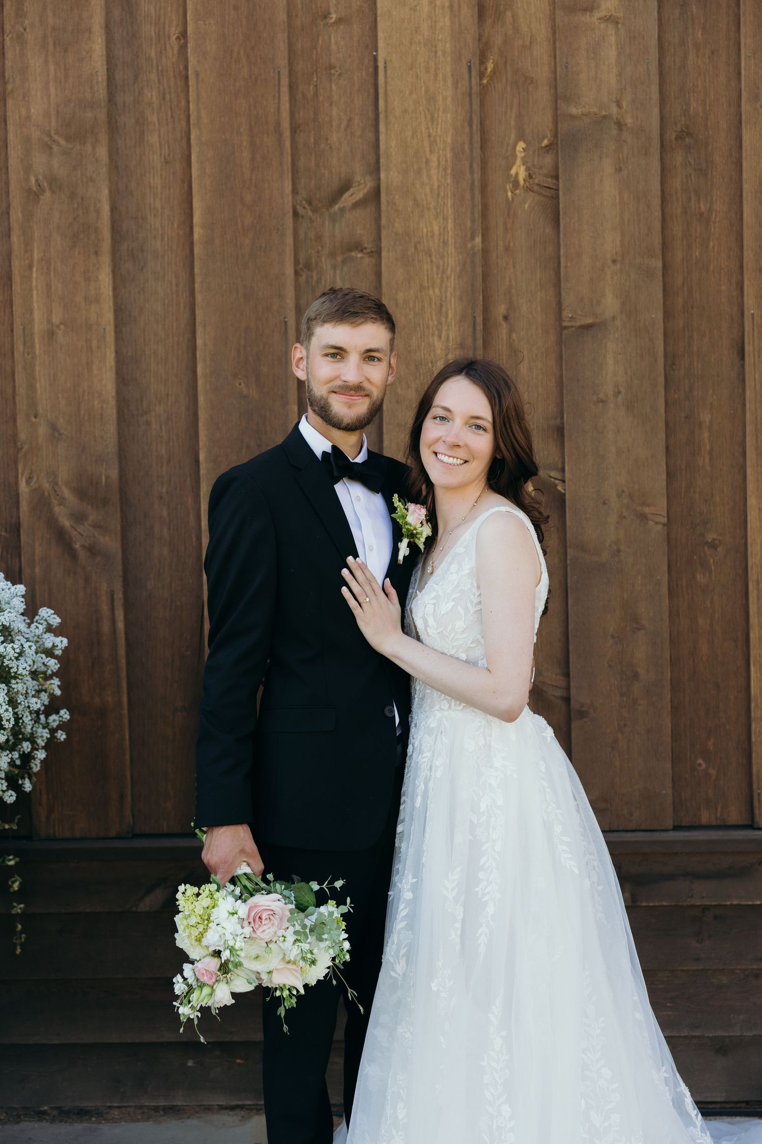 A wedding couple poses against a rustic wooden wall decorated with white floral arrangements.