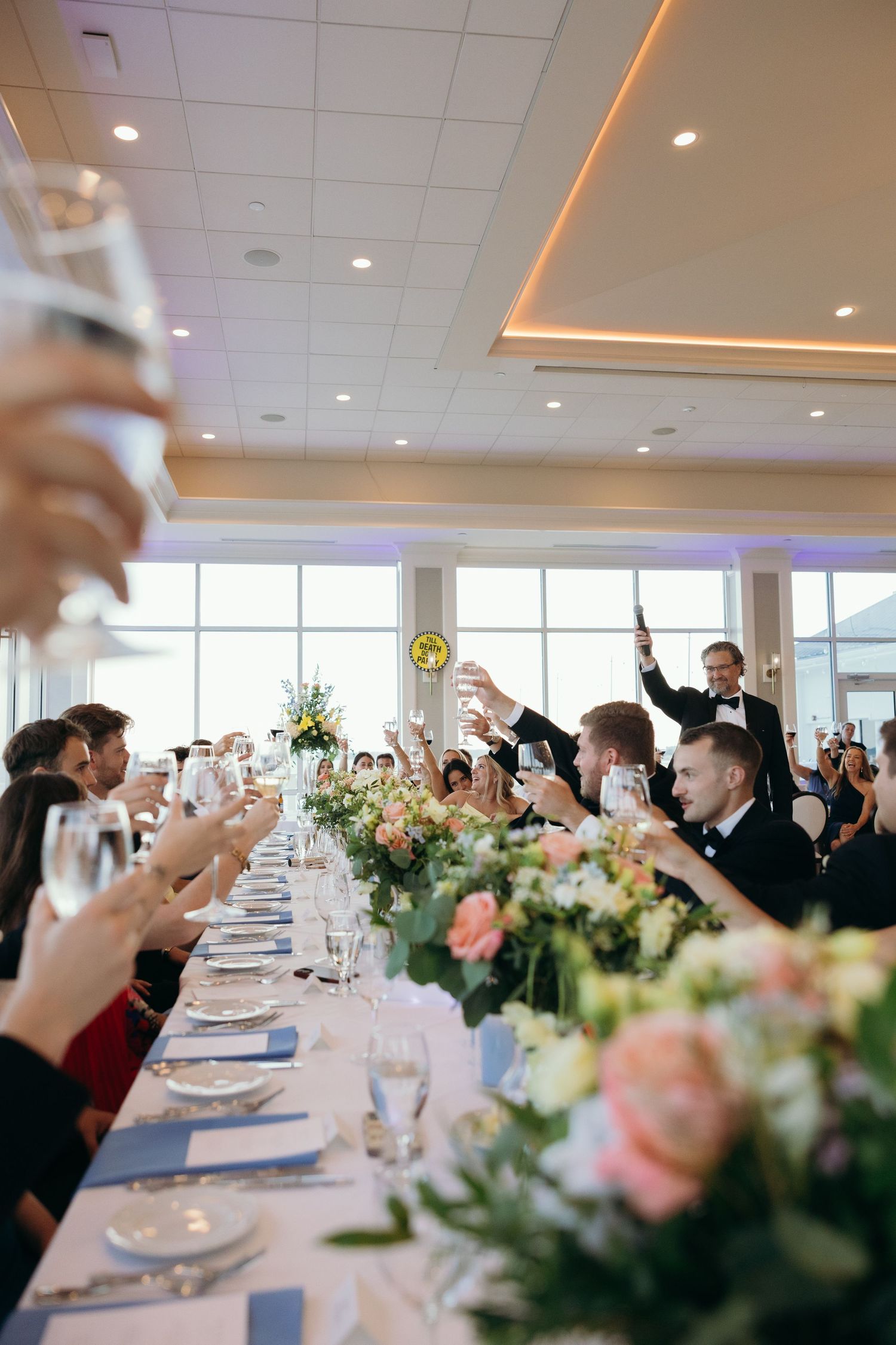 Wedding reception guests raising glasses in a celebratory toast at decorated dining tables.