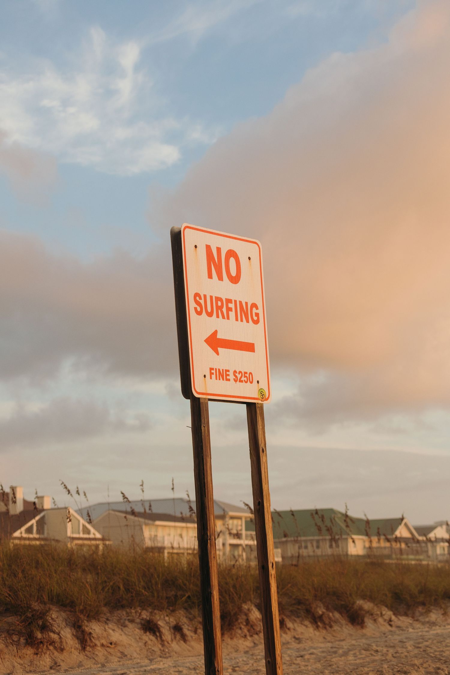 A 'No Surfing' warning sign stands on a beach with coastal homes and cloudy sunset skies in the background.