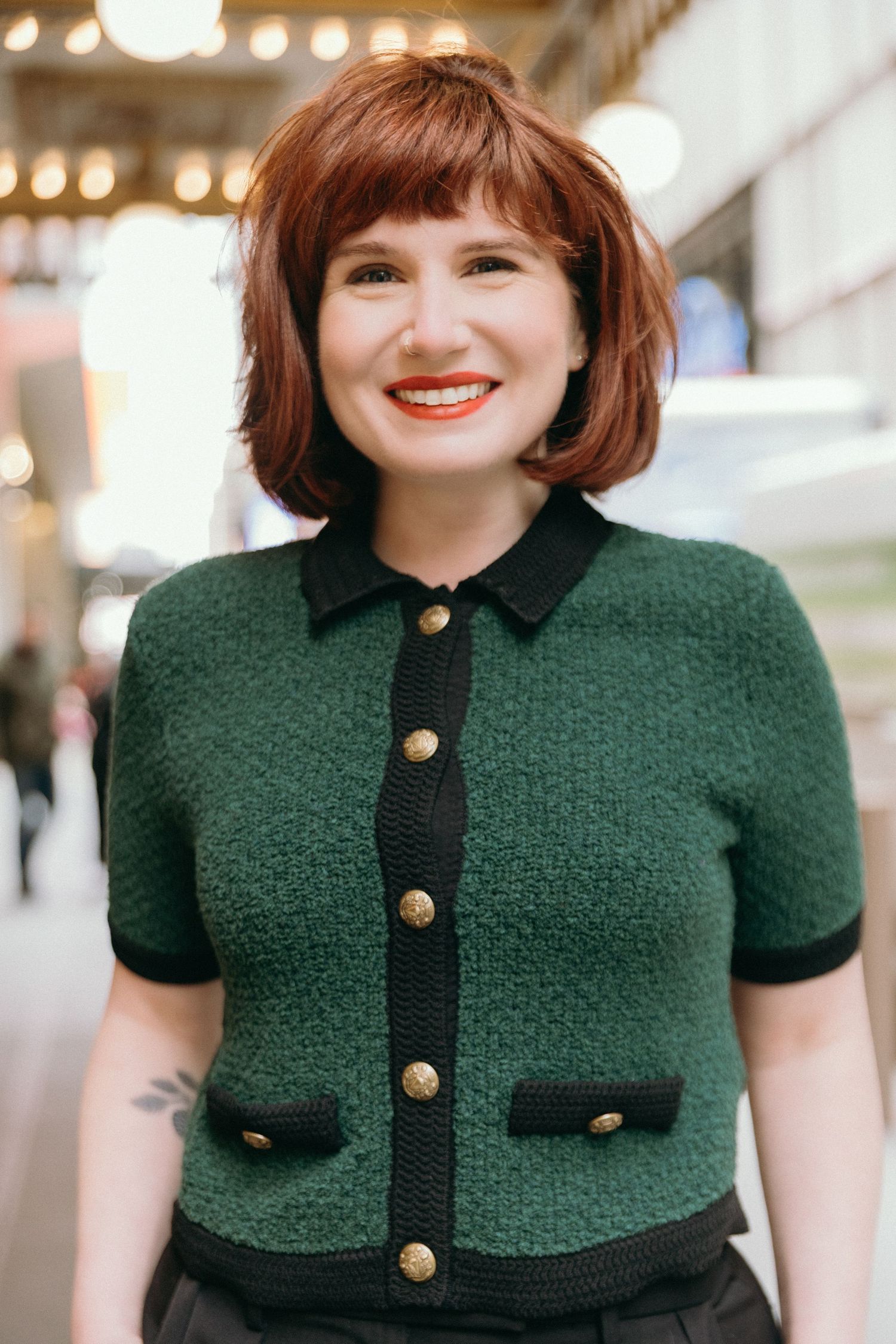A woman wearing a dark green tweed jacket with black trim and gold buttons poses in a bright indoor setting.
