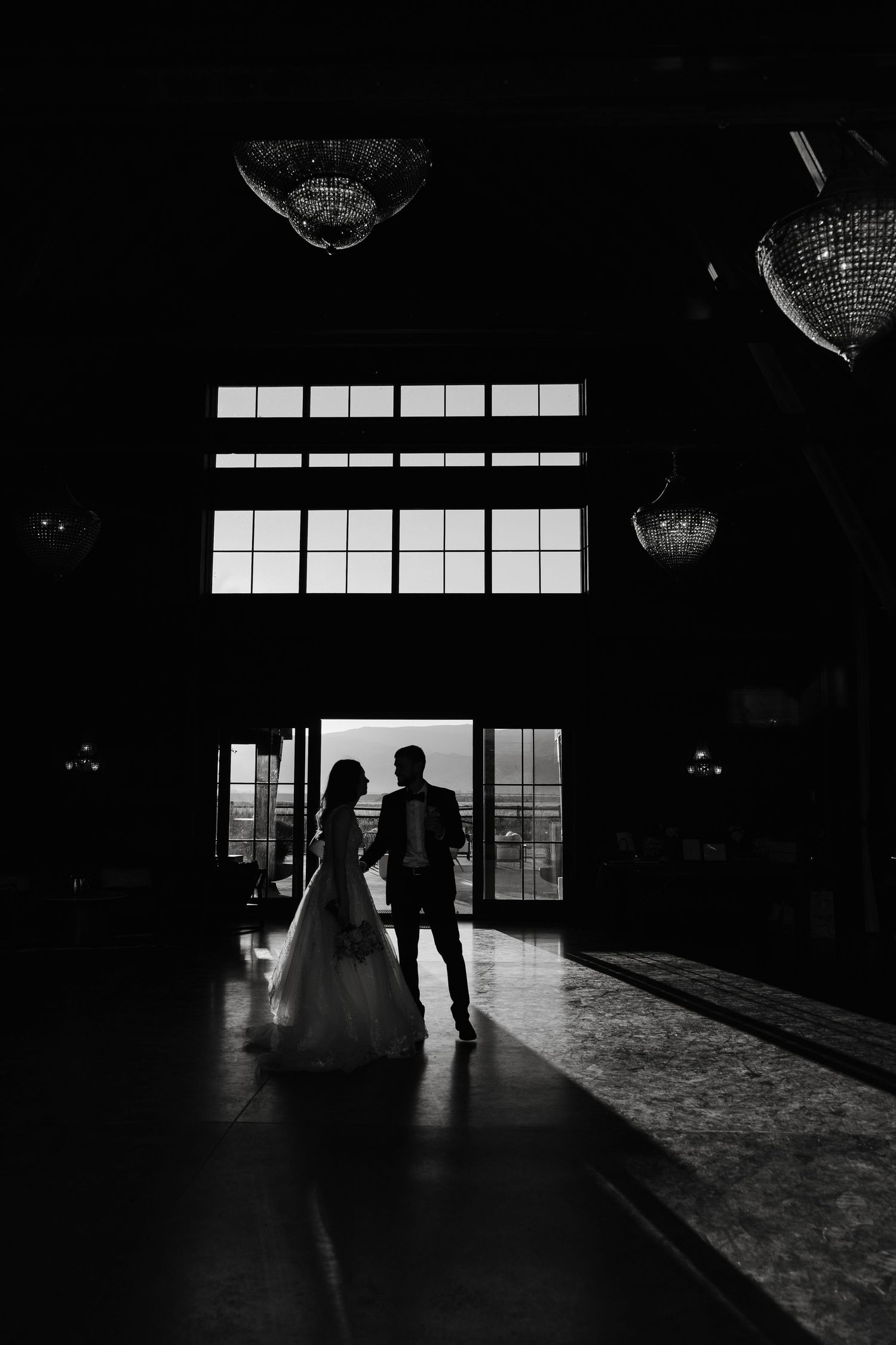 Silhouettes of wedding couple in dark indoor venue with window light.