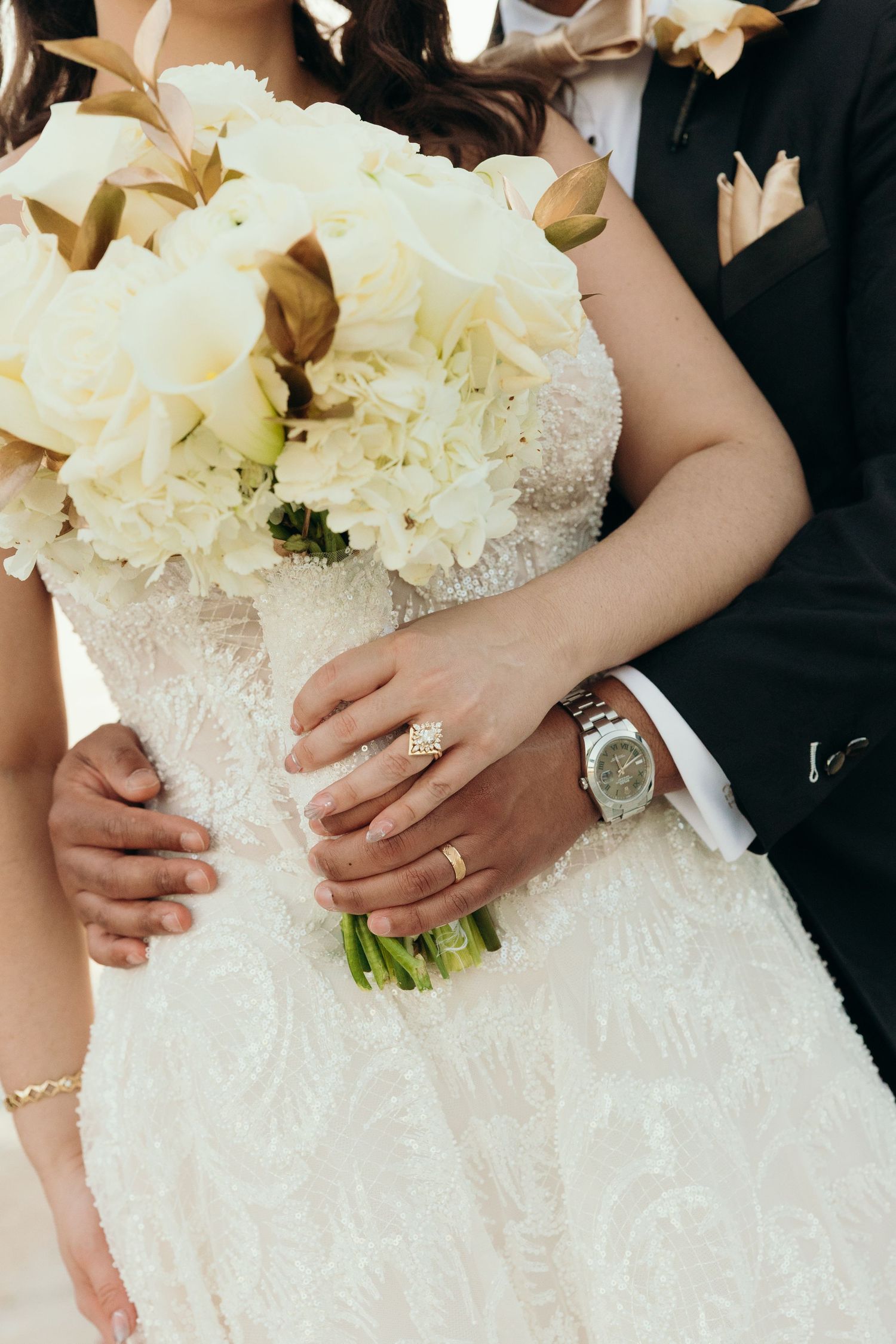 A close-up of a white floral bouquet held against a lace wedding dress with jewelry details visible.