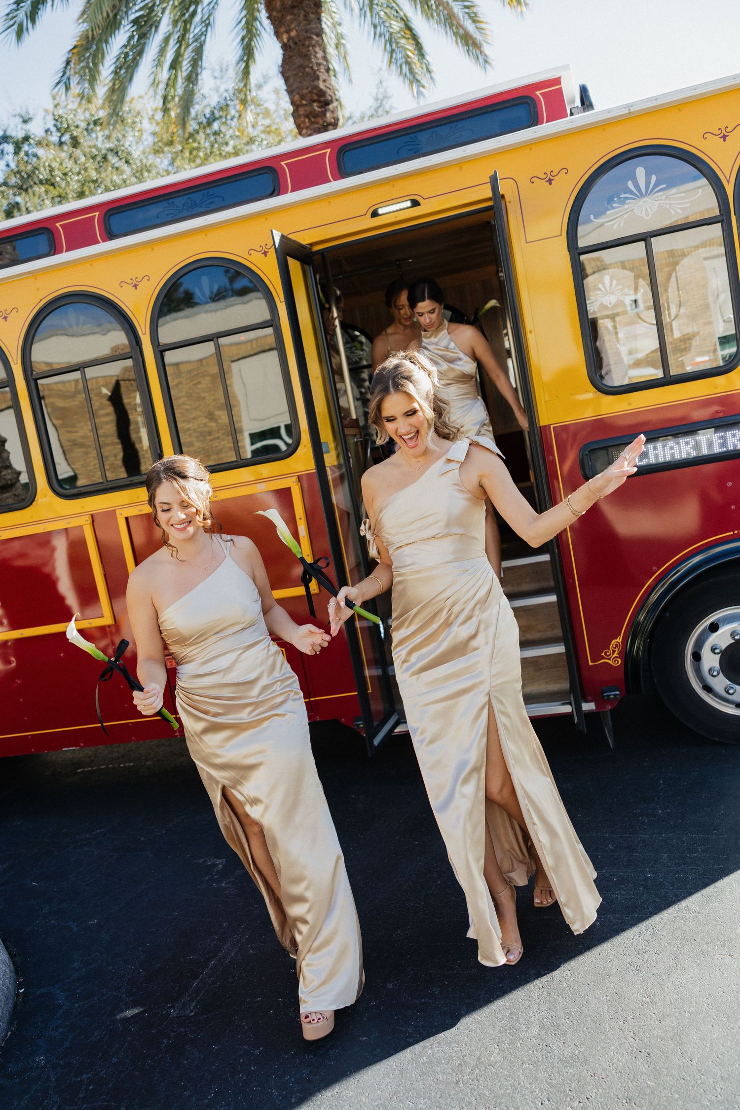 Wedding party members in champagne-colored dresses exit a vintage red and yellow trolley bus under palm trees.