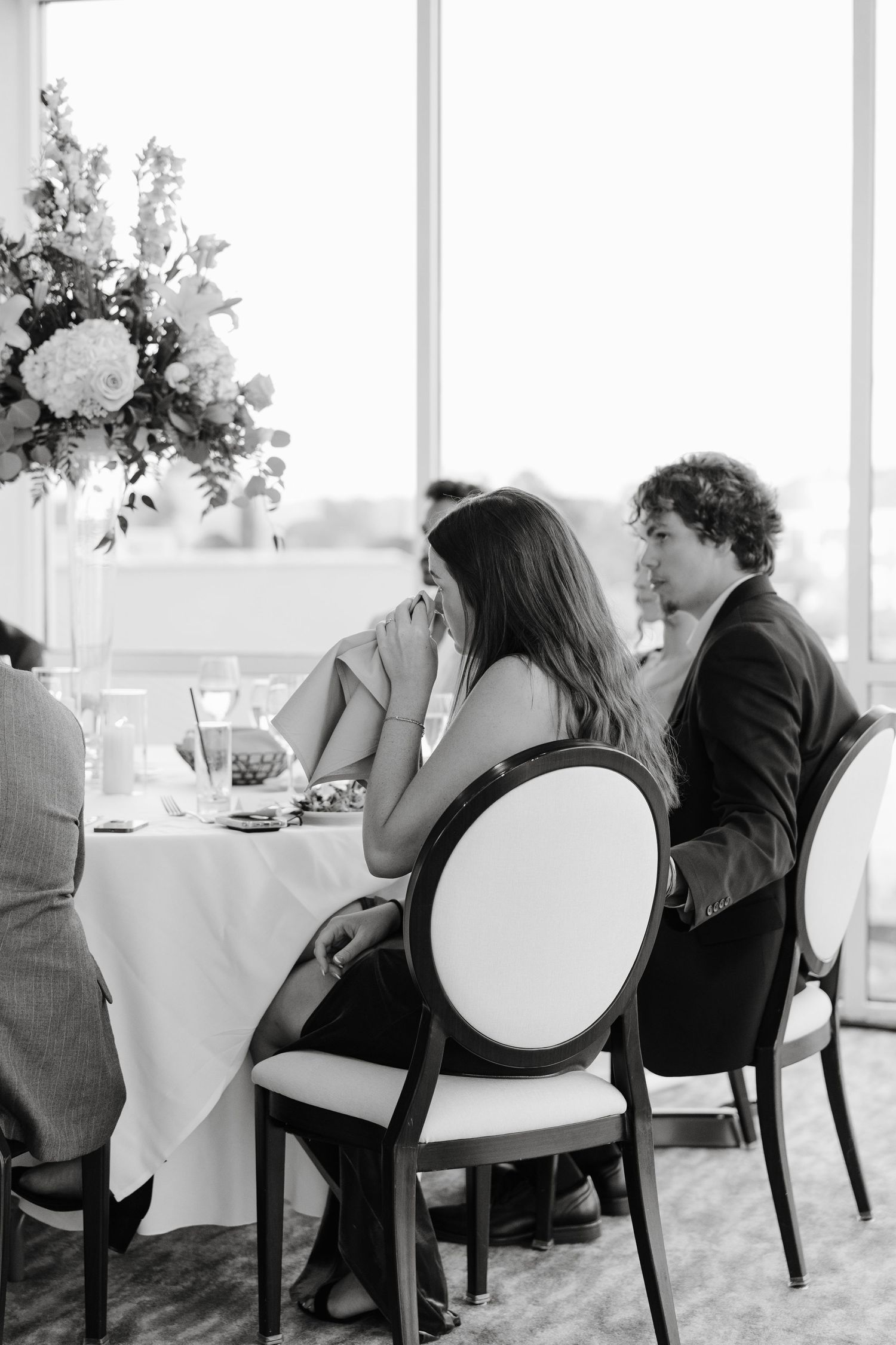 Elegant dining setup with white tablecloths and floral centerpieces in a bright room with large windows.