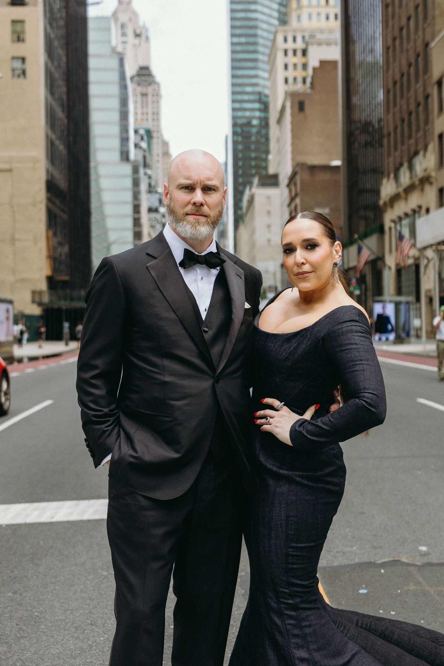 A couple in formal black attire poses on an empty city street with skyscrapers in the background.