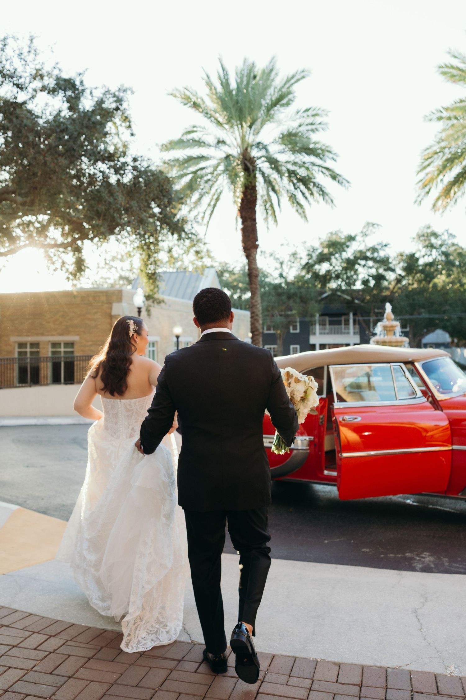 A couple in wedding attire walks together near palm trees with a classic red vintage car in the background.