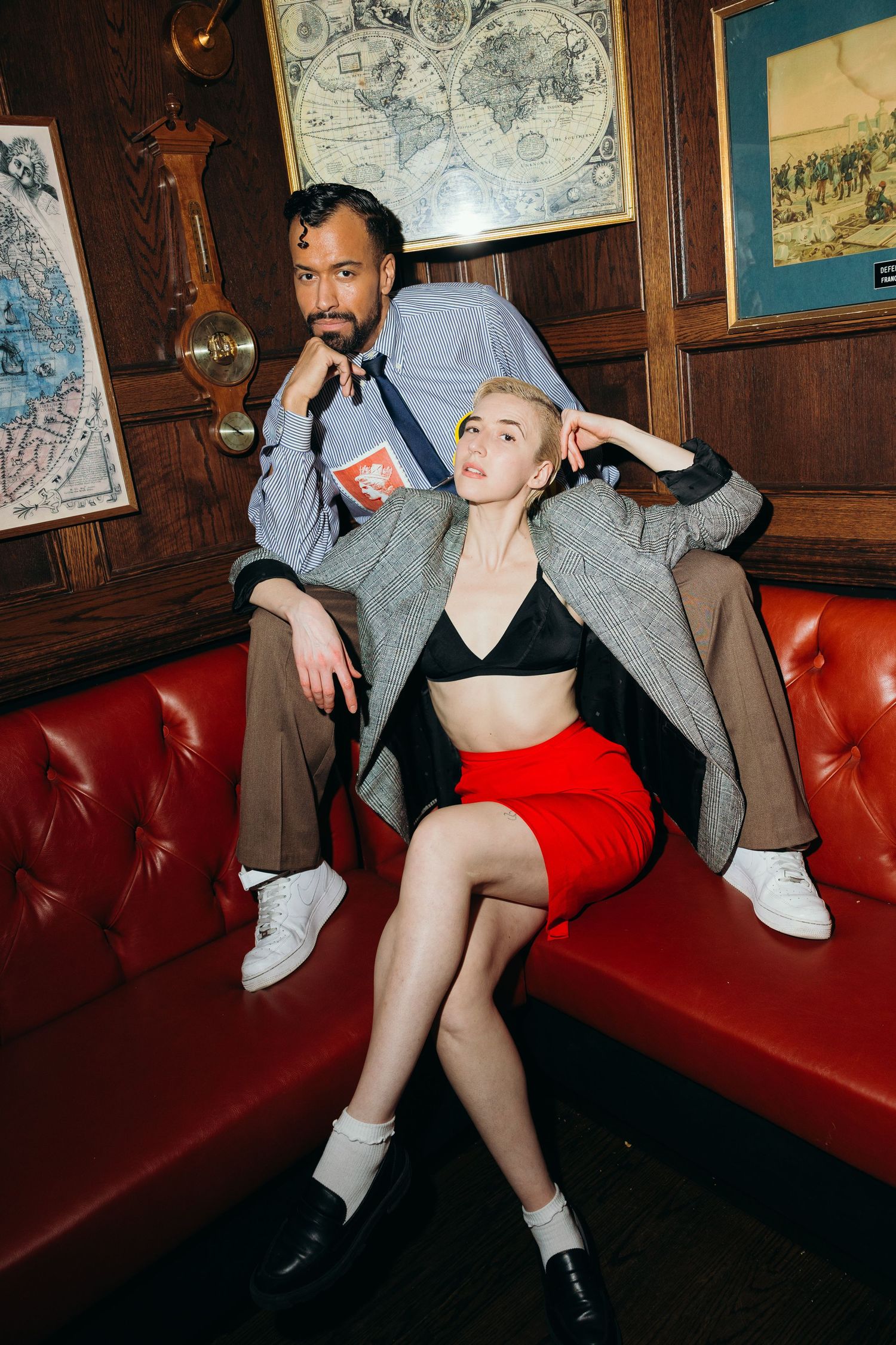 Man and woman posing on red leather sofa in wood-paneled bar, she wears red skirt and black bralette.