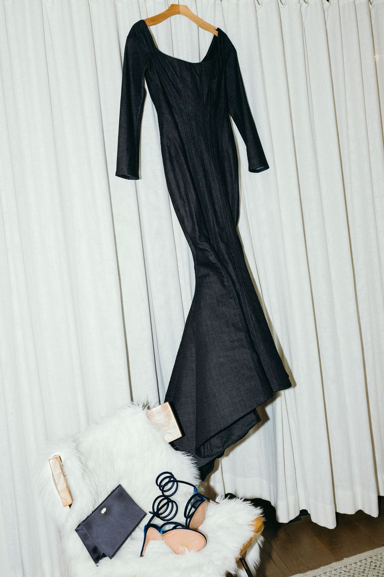 Black dress hanging on wooden hanger against white curtained background with items scattered below.