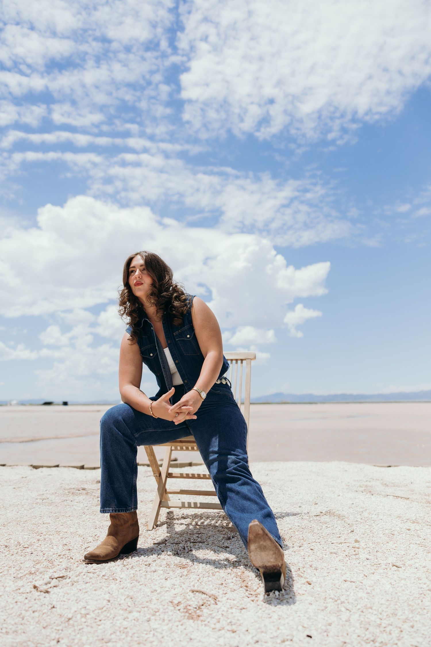 Person in denim sits contemplatively on a wooden chair against stark white salt flats under cloudy blue skies.