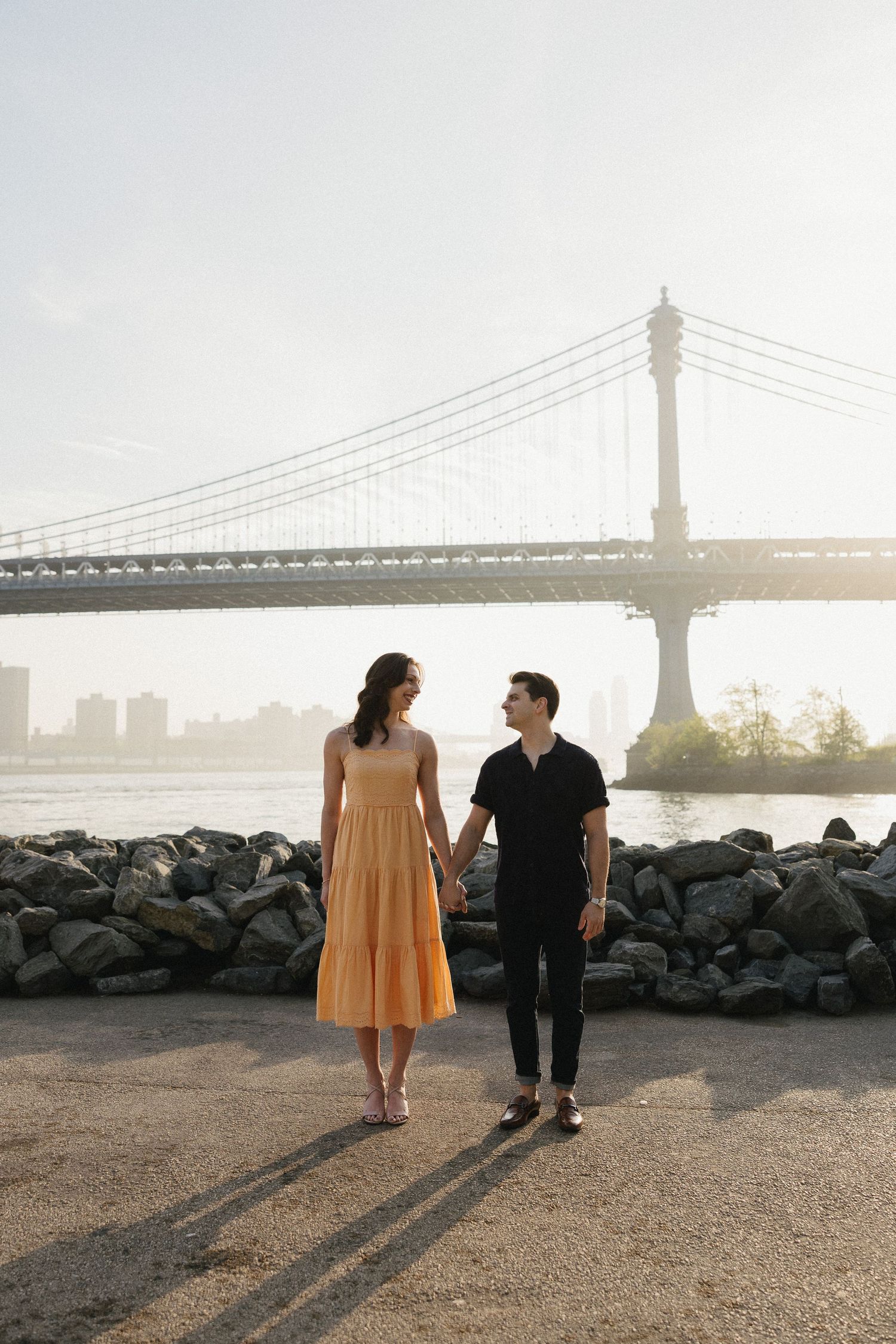 A couple stands together on a rocky shore with a bridge silhouetted against the bright sky at sunset.
