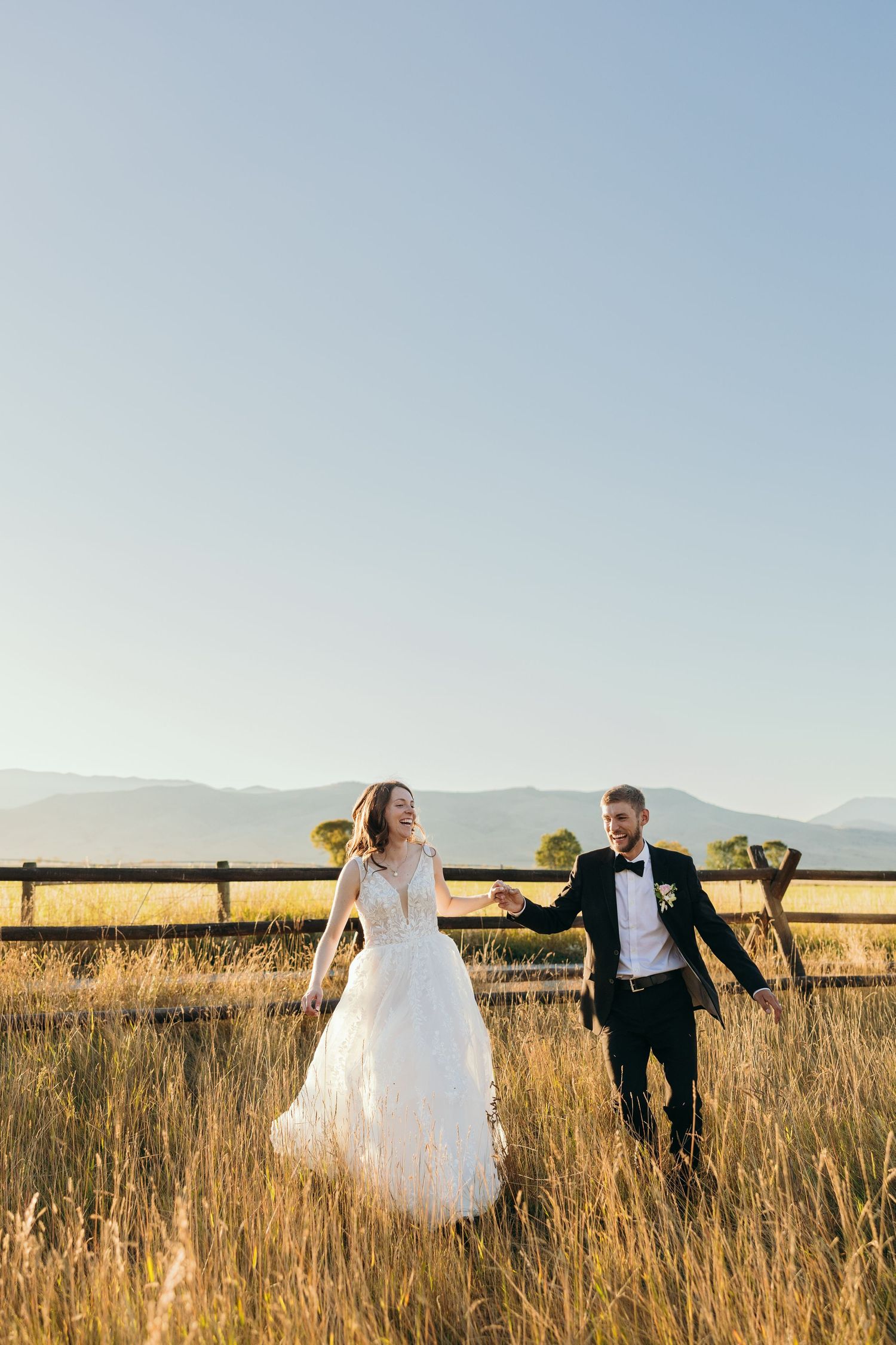 A couple runs joyfully through a golden field with mountains in the background on their wedding day.