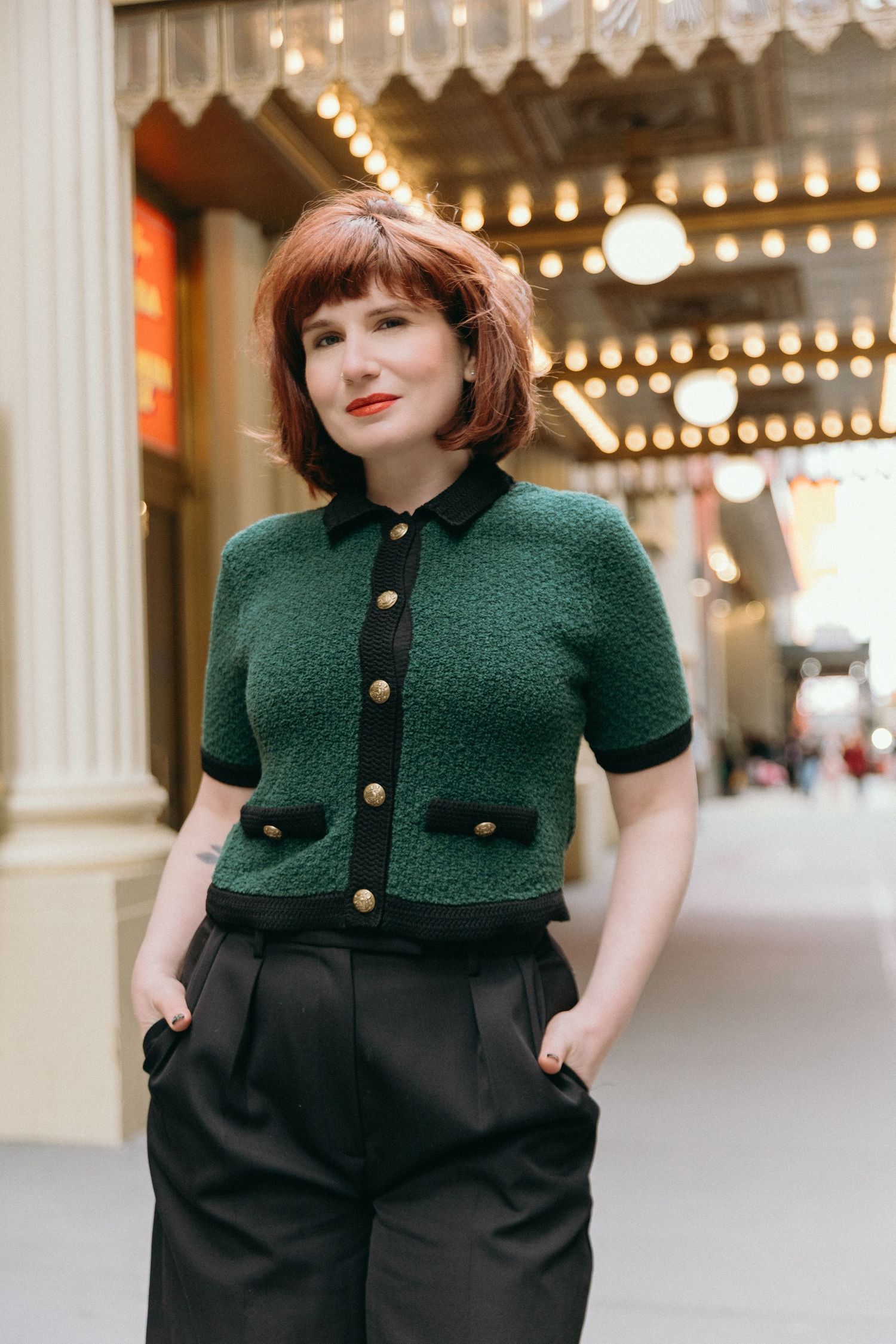 A vintage-inspired outfit featuring a dark green knit cardigan paired with high-waisted black trousers and retro lighting.