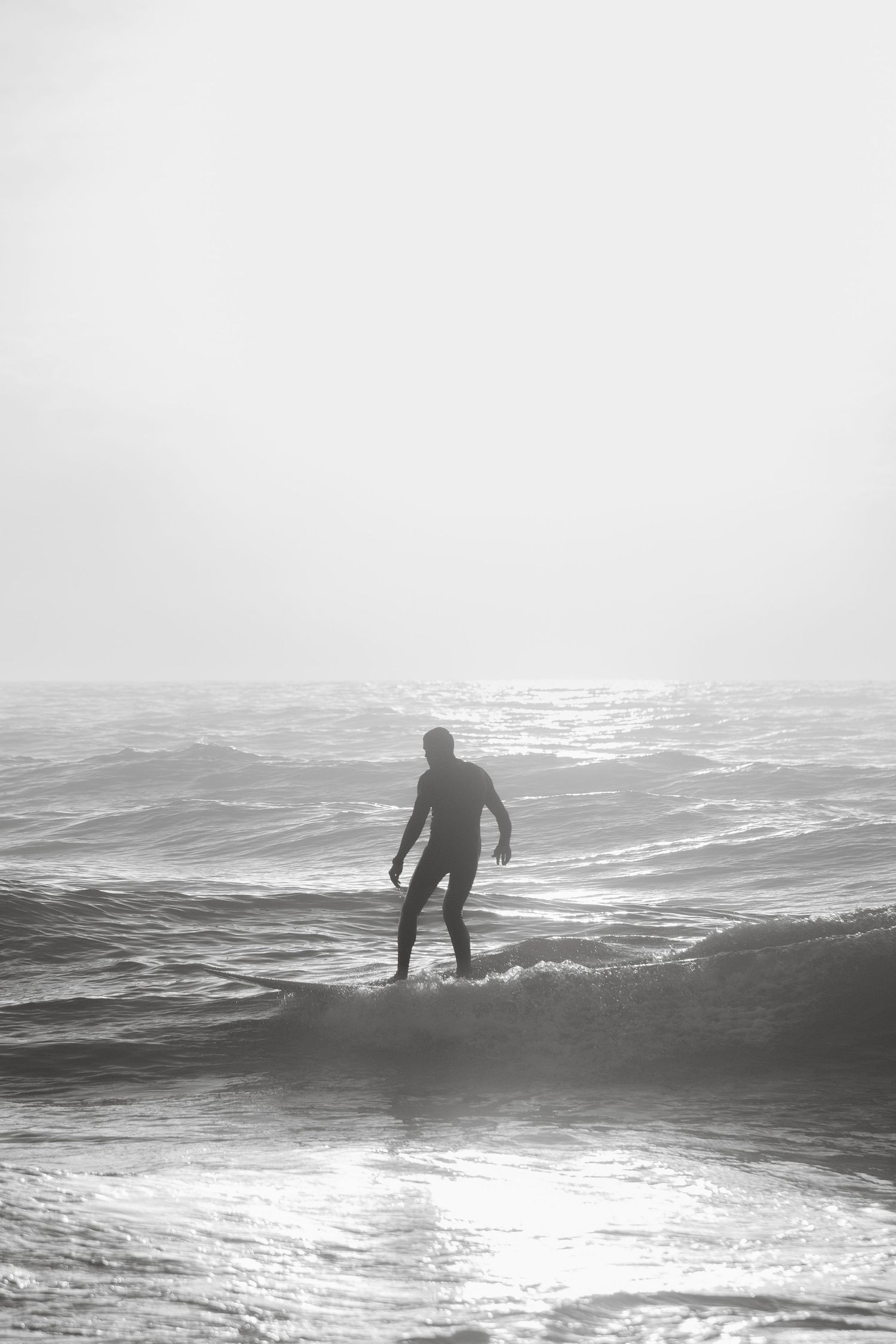 Silhouetted surfer rides a wave against a bright ocean horizon in black and white.