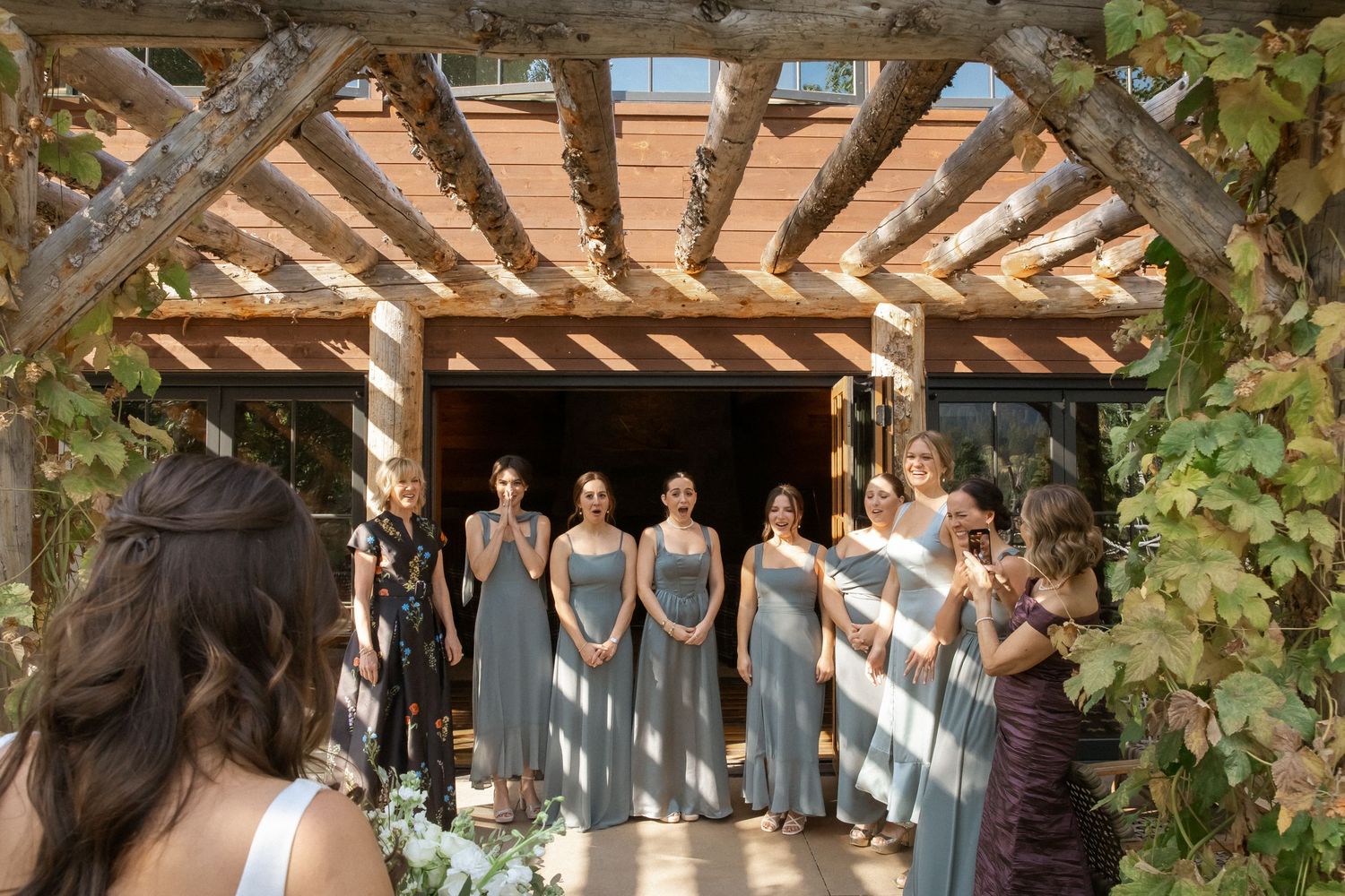 Wedding party members stand in a row under a rustic wooden pergola decorated with green vines and foliage.