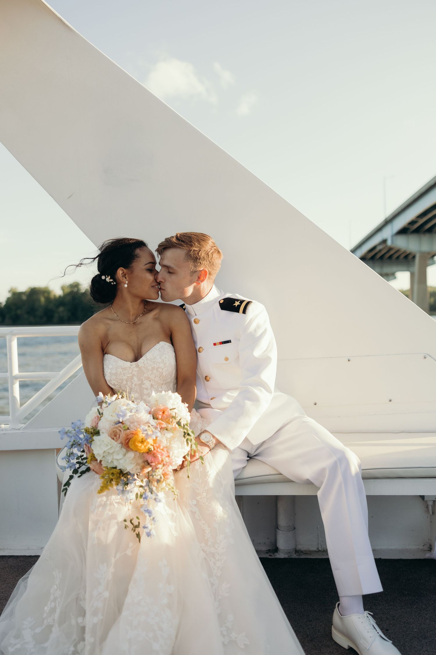 A romantic wedding couple shares a kiss on a white deck, with the bride holding a peach and white floral bouquet.